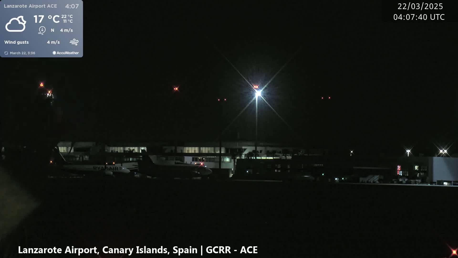 At night, under a partly cloudy sky with a light north wind, several airplanes are parked on a dark airport tarmac illuminated by bright lights.