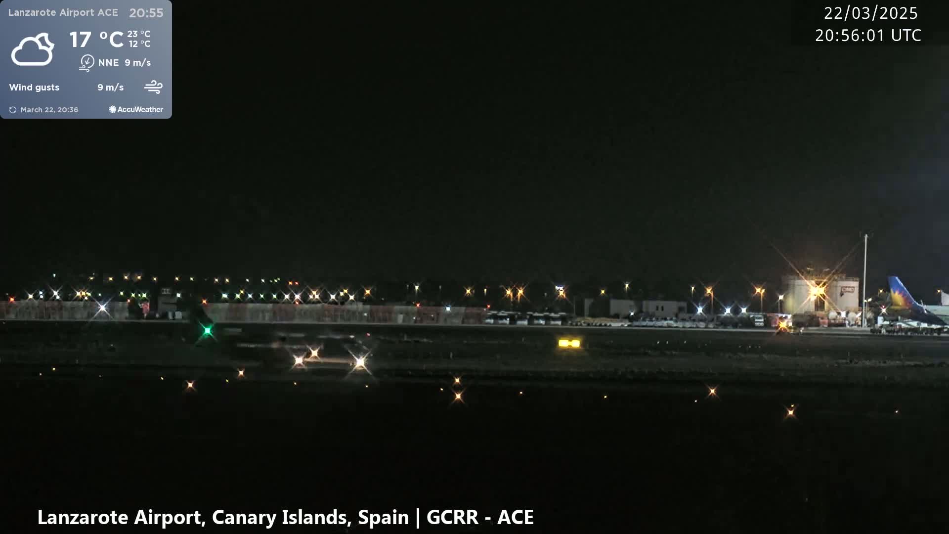 A nighttime view of an airport runway with lights illuminating the tarmac and surrounding buildings under a partly cloudy sky.