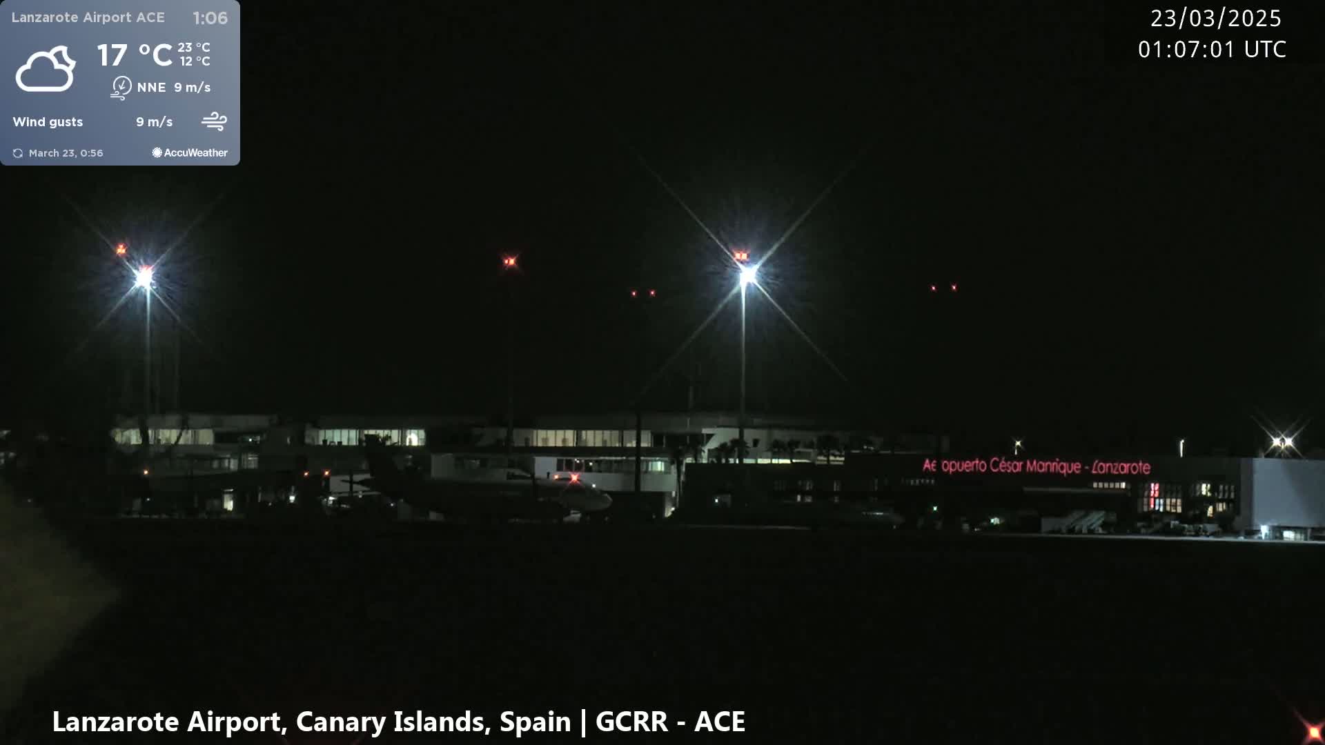 A nighttime view of an airport terminal with several bright lights and some aircraft on the tarmac under partly cloudy skies.