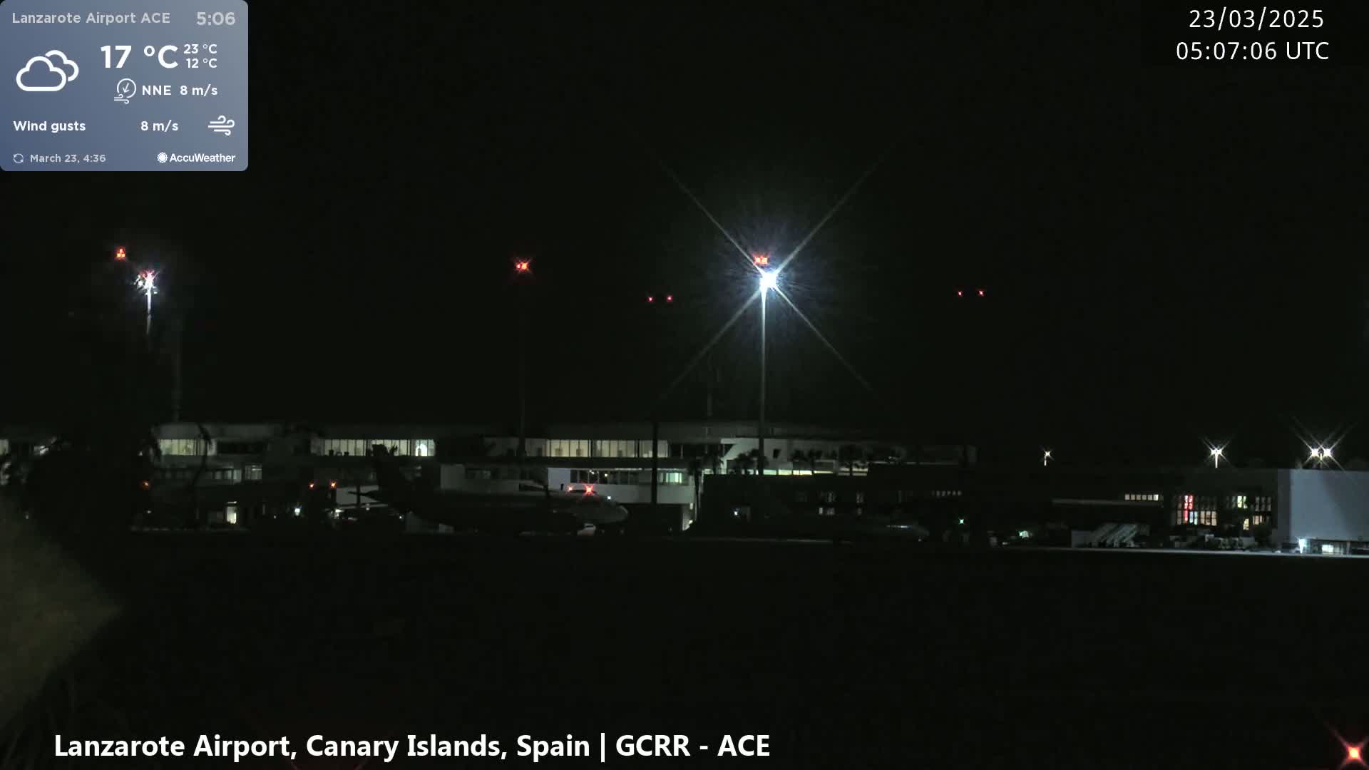 A nighttime view of an airport with a plane on the tarmac, illuminated by bright lights against a dark, cloudy sky.