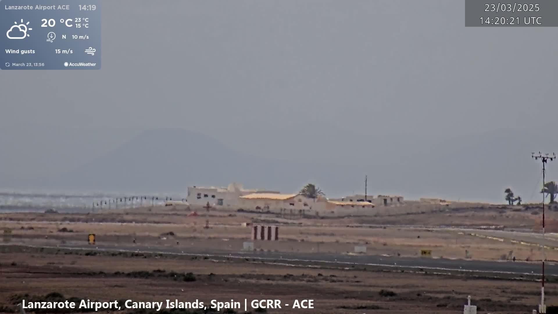 A hazy, partly sunny day shows a low-lying building complex near a runway, with a distant mountain range visible under an overcast sky.