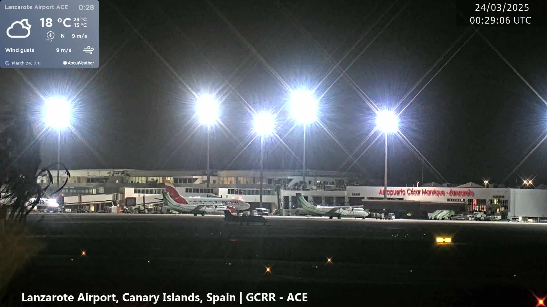 At night, under a partly cloudy sky with a northerly wind, two airplanes are parked on the tarmac of a brightly lit airport.