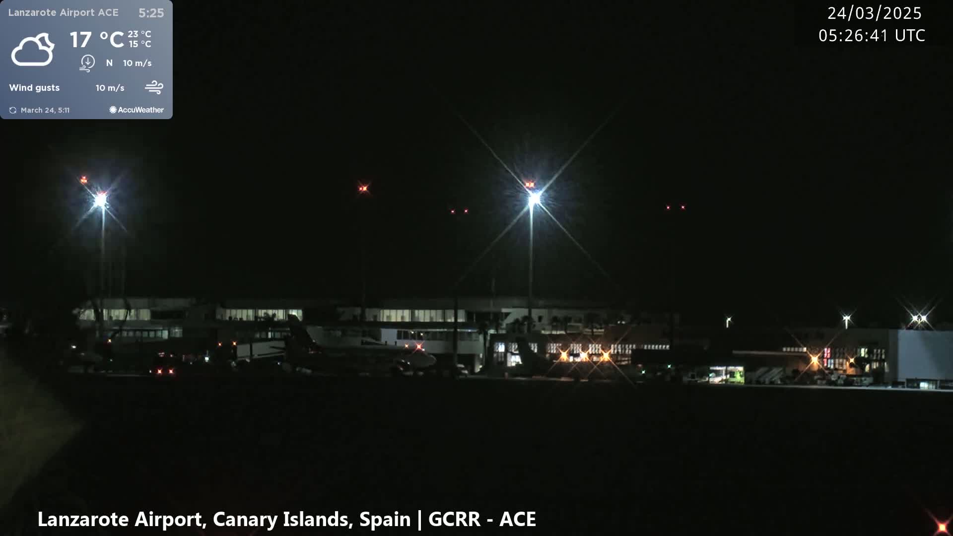 A nighttime view of an airport with several planes on the tarmac, illuminated by bright lights under a clear, dark sky with a slight wind.