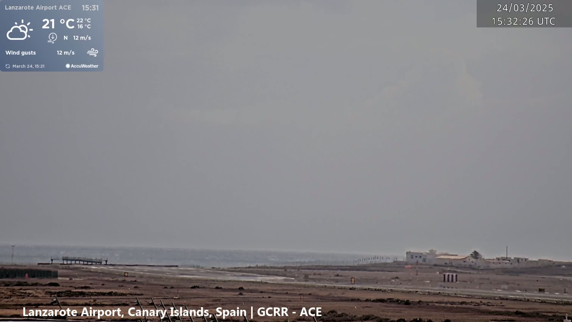 A hazy, overcast day at an airport shows a runway, buildings, and the ocean in the distance, with a light wind.