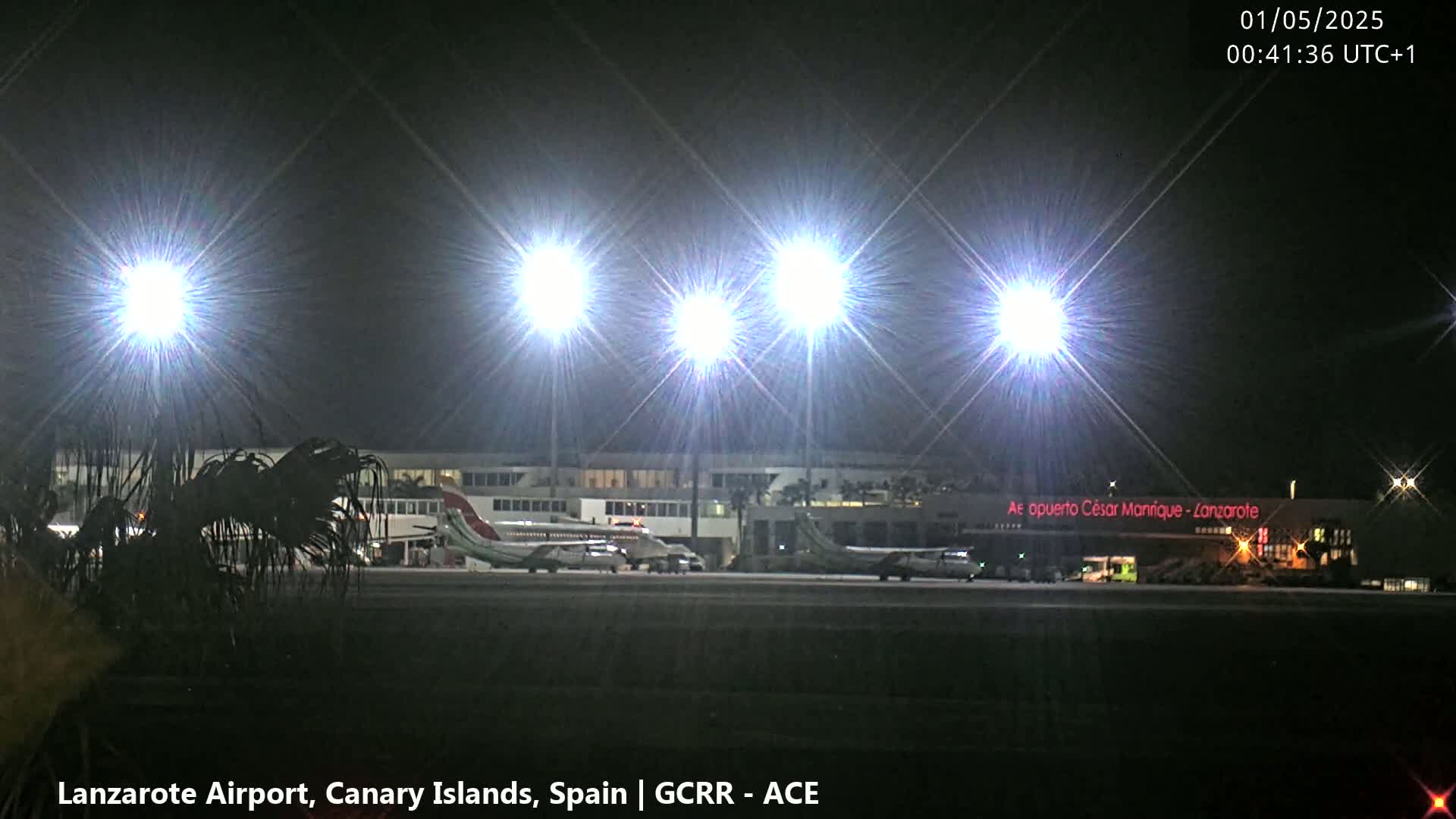 At night, bright floodlights illuminate two airplanes parked on a tarmac in front of an airport terminal building.