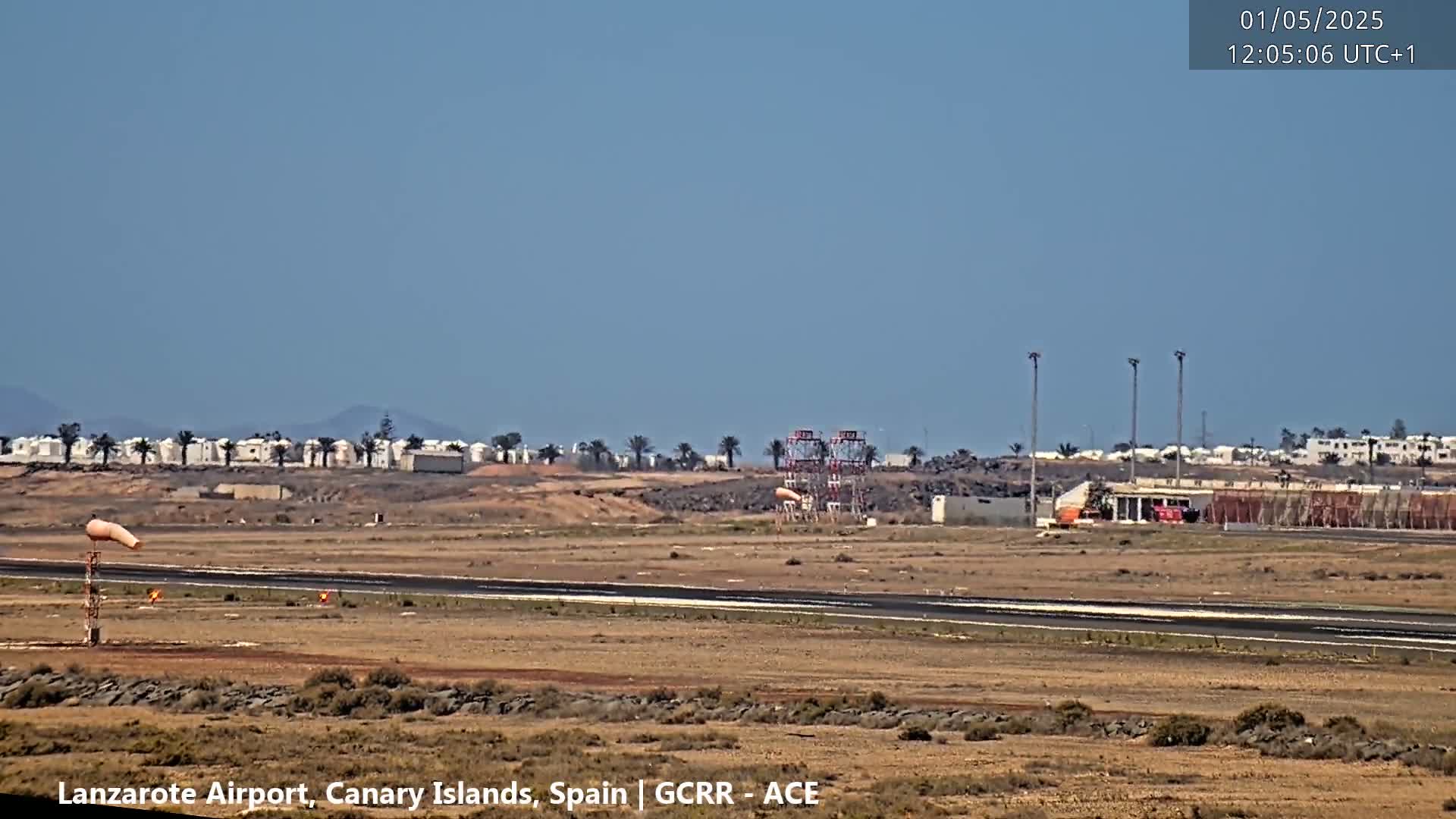 A windsock and airport runway are visible on a clear, sunny day, with buildings and palm trees in the distance.