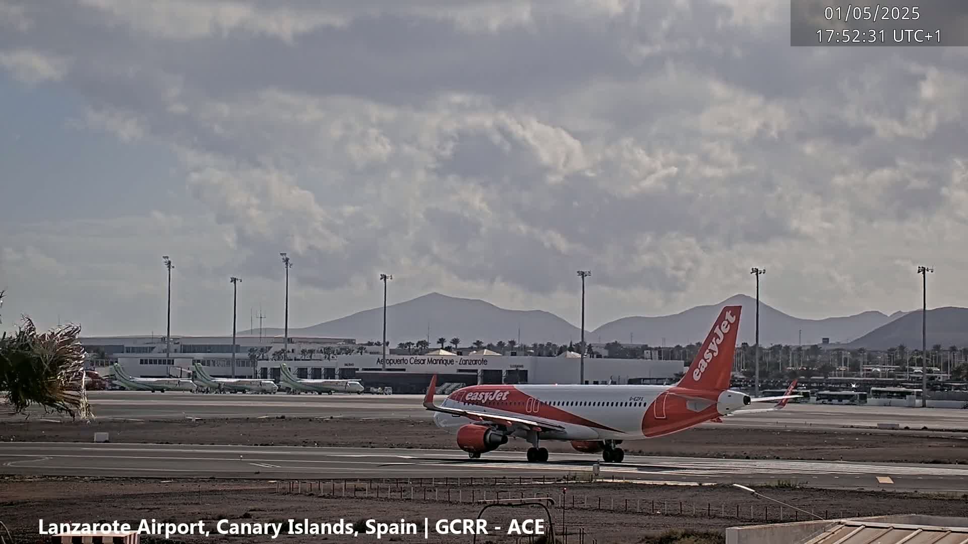 An easyJet airplane taxis on an airport tarmac under a partly cloudy sky, with mountains in the background.