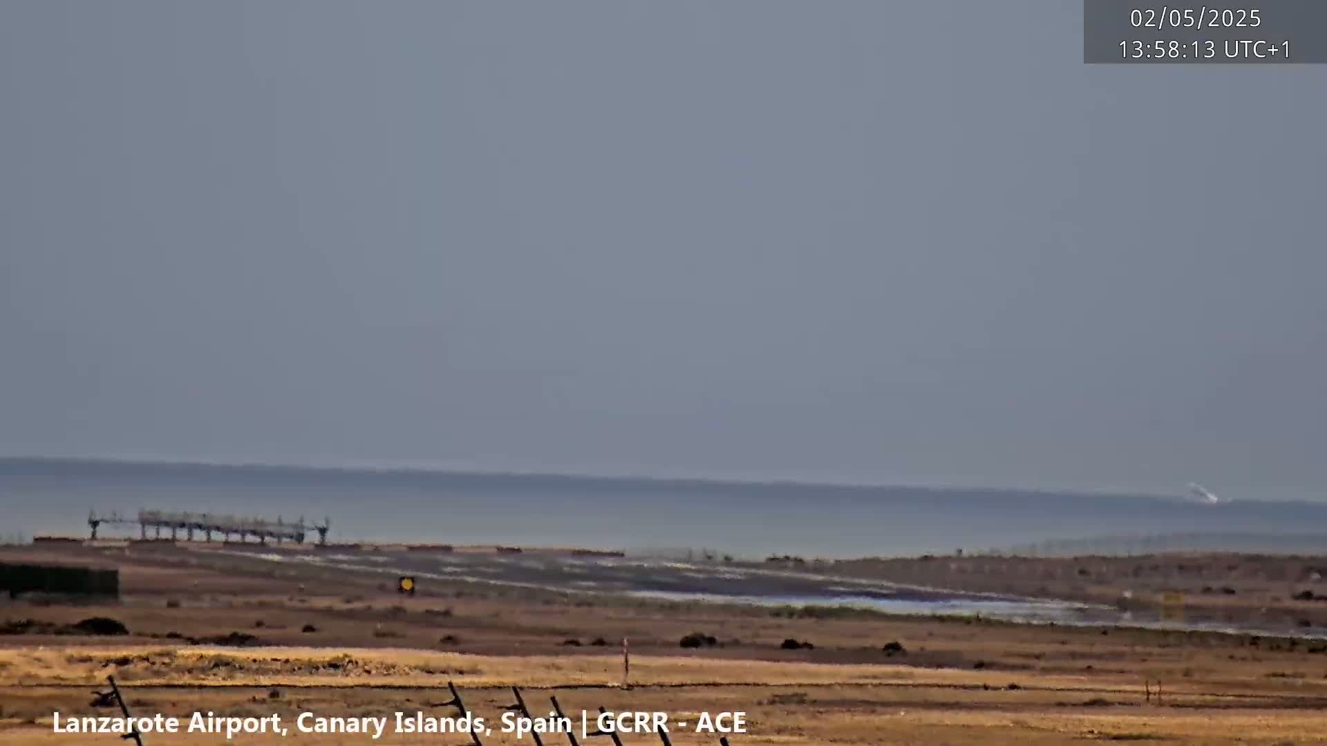 A flat, dry, brown landscape extends to a calm ocean under a hazy sky.