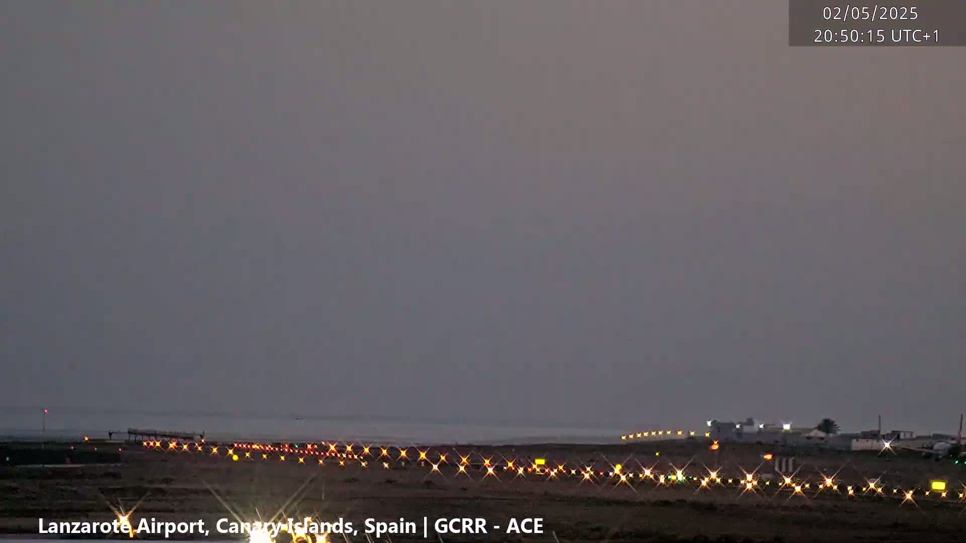 Under a dusky sky, runway lights illuminate a dark airport tarmac next to a calm body of water.