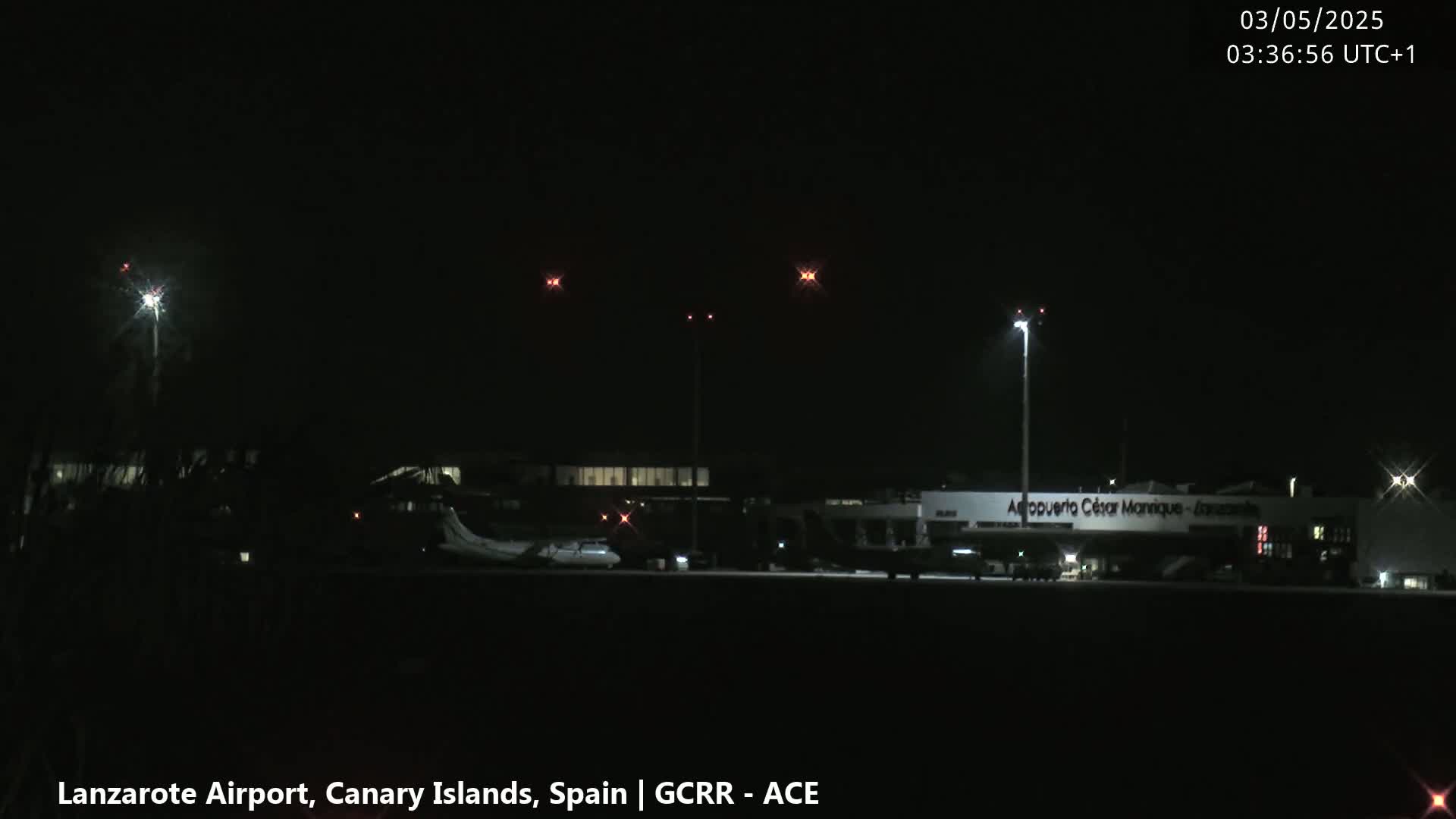At night, a plane sits on a runway at an airport with several lights illuminating the tarmac and terminal buildings under a dark sky.