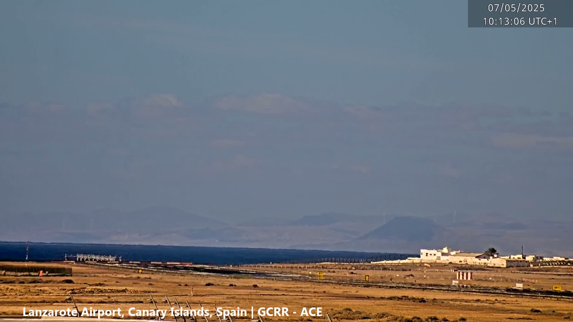 A hazy, sunny day reveals a flat, arid landscape with a distant ocean and mountains, and airport infrastructure in the foreground.