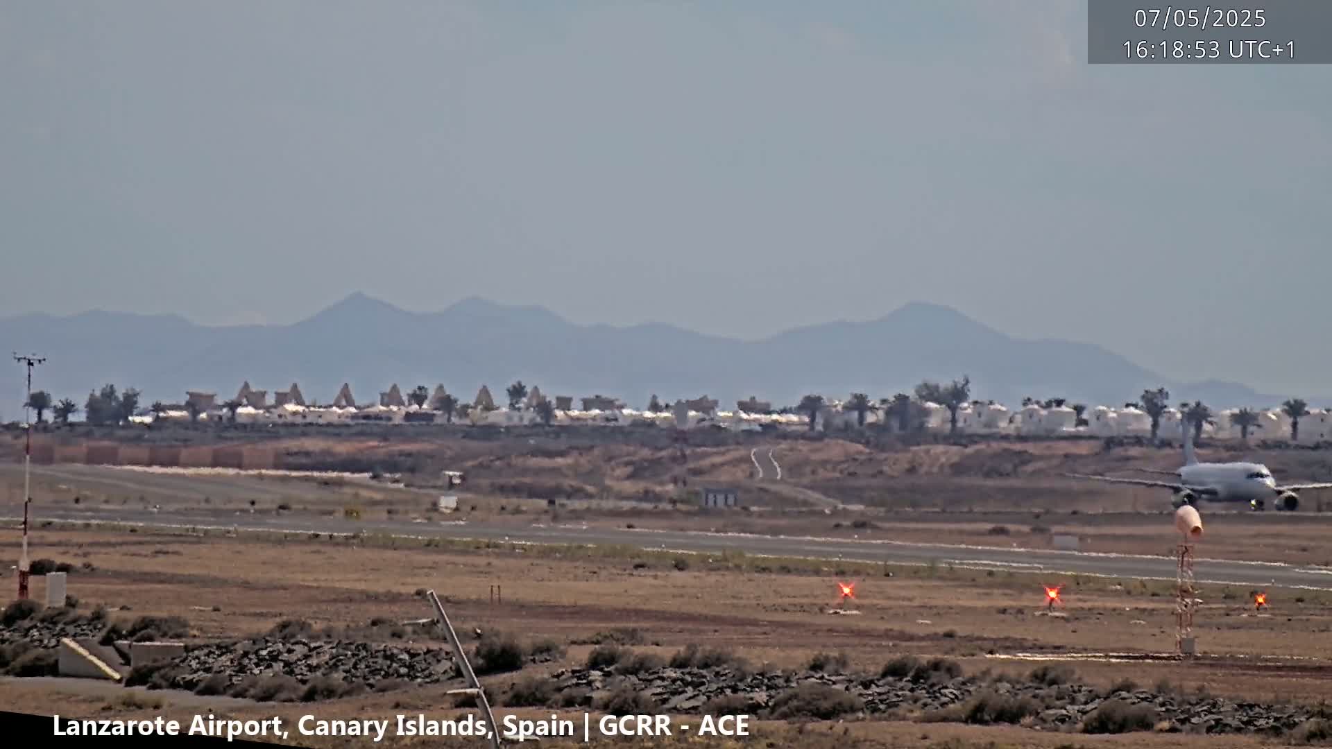 A plane taxis on a runway near a low-lying building complex with distant hazy mountains under a clear sky.
