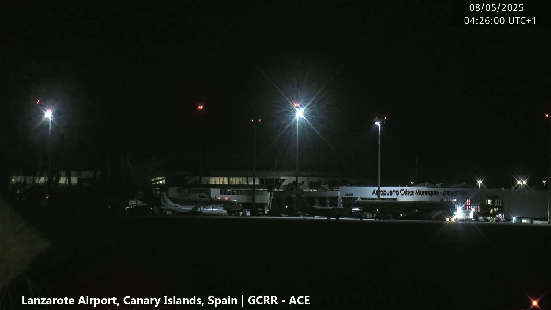 A nighttime view of an airport terminal building and several airplanes on the tarmac, illuminated by bright lights under a clear, dark sky.