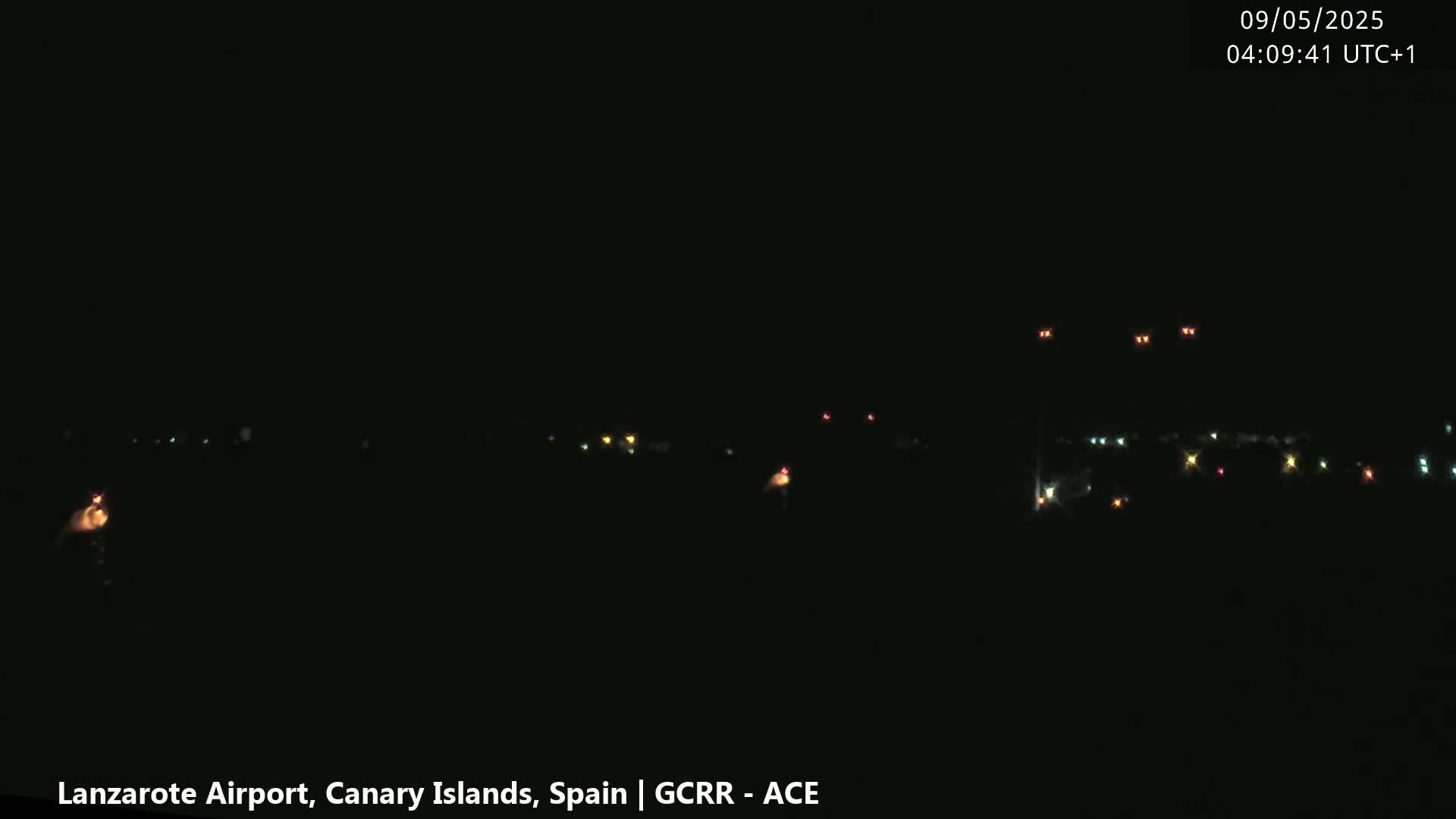 A nighttime view of a dark airport runway with scattered lights visible in the distance.