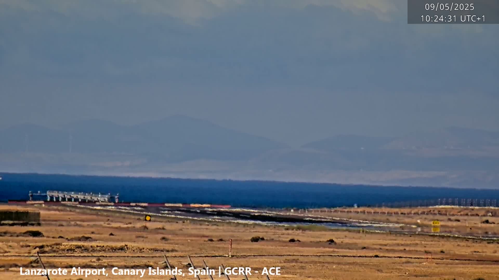 A hazy, distant view of mountains and ocean, with airport infrastructure visible in the foreground under a clear sky.