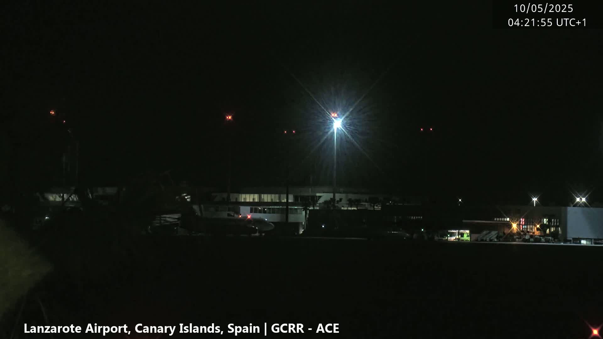 A brightly lit airport at night is shown with an airplane on the tarmac, several distant lights, and a dark sky.