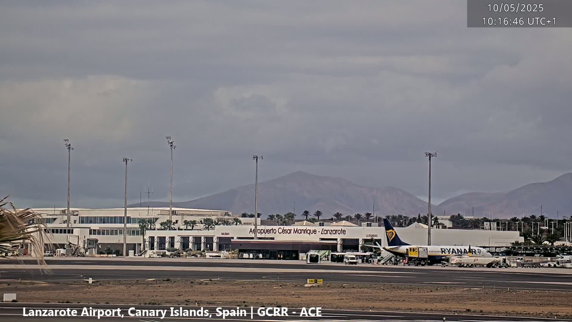 An airport terminal with a Ryanair plane parked at the gate, under an overcast sky, and mountains in the background.