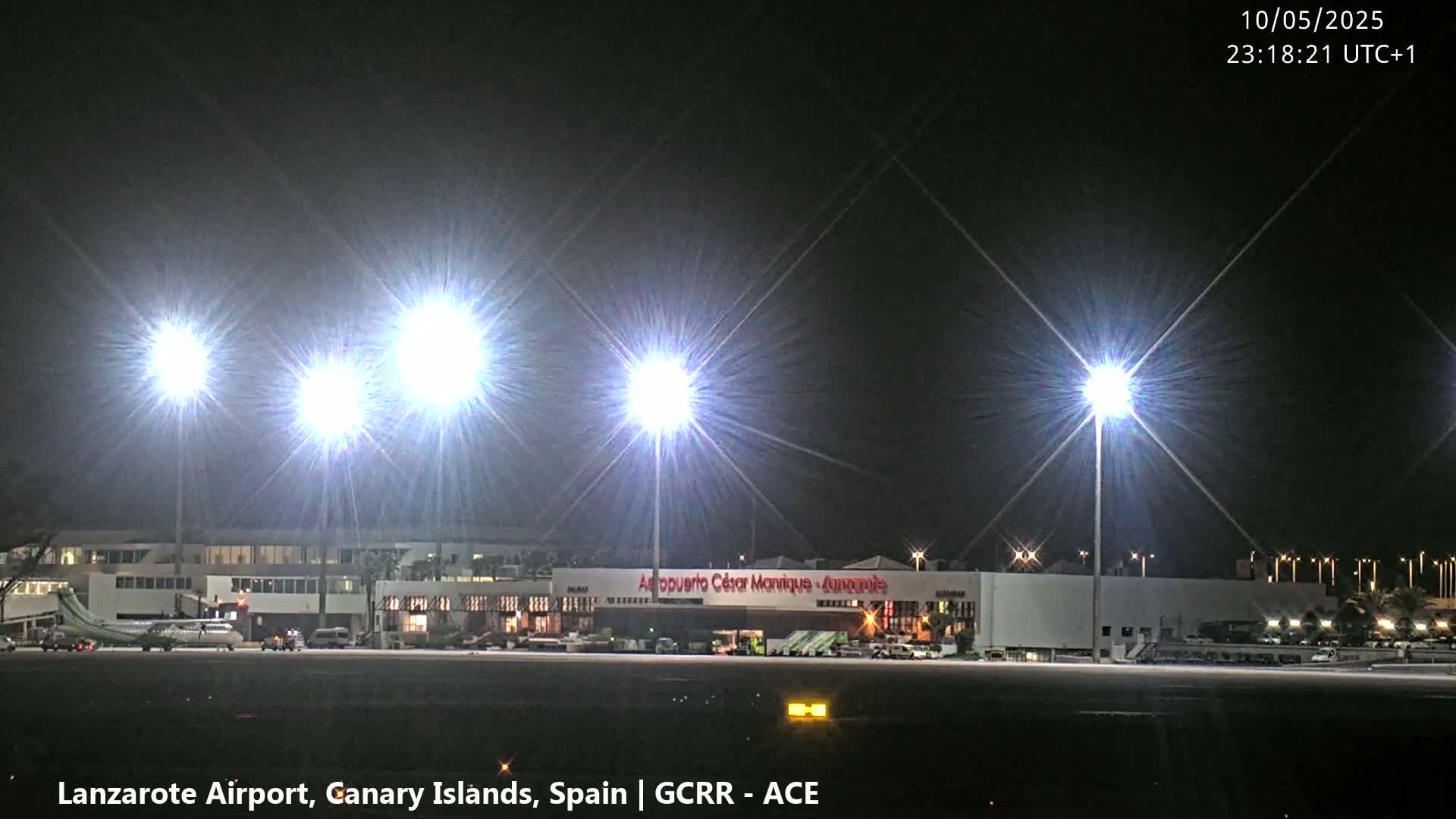 At night, brightly lit airport buildings and tarmac are visible, with airplanes parked nearby under clear skies.