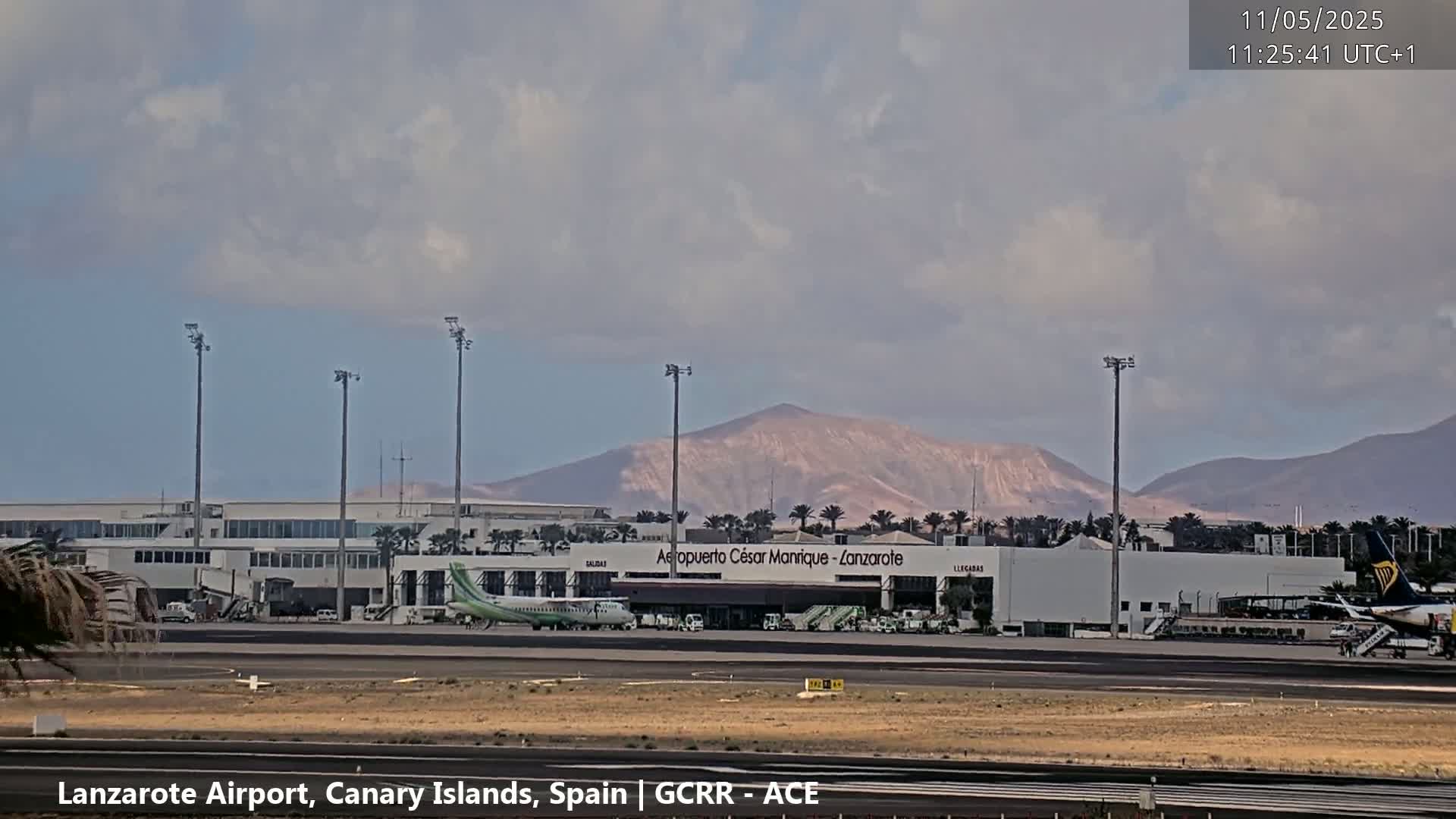 An airport terminal building with several planes on the tarmac is situated in front of a mountainous landscape under a partly cloudy sky.