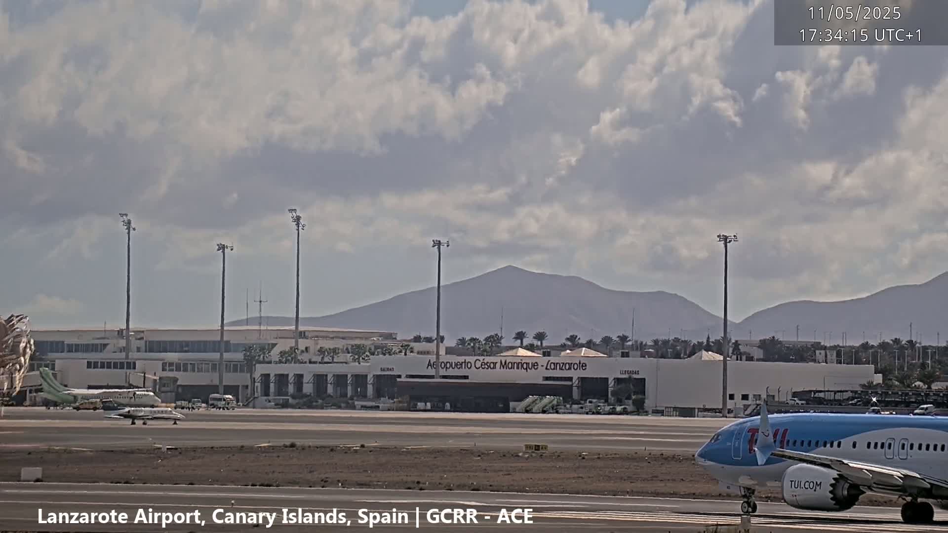 A partly cloudy day at an airport shows several planes on the tarmac, with mountains in the background.