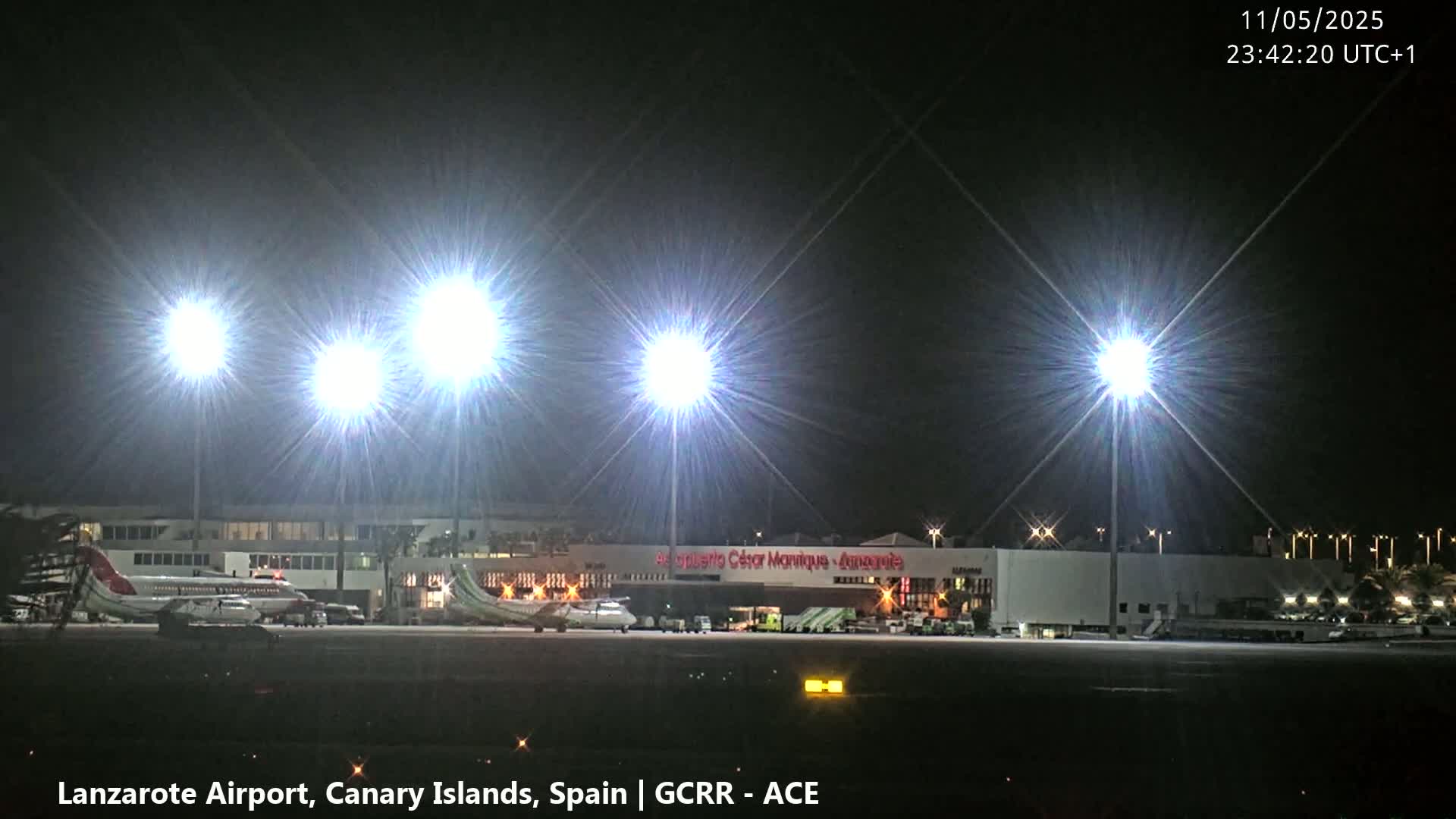 At night, brightly lit airplanes are parked on a tarmac in front of a brightly lit airport terminal building.