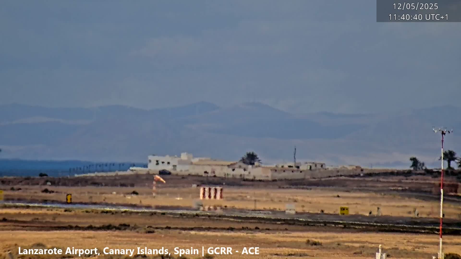 A flat, dry landscape with low buildings and a windsock is visible under a hazy sky, with distant mountains in the background.