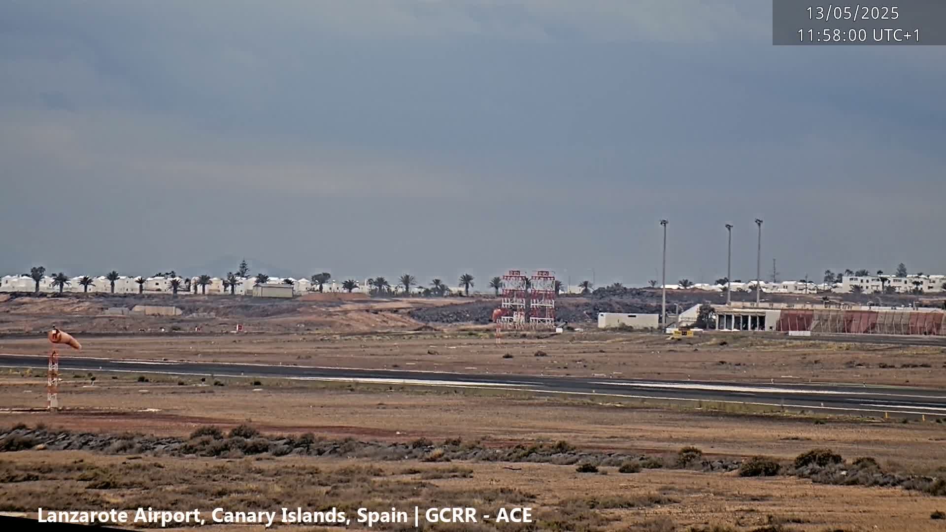 A flat, arid landscape with a runway, airport buildings, and distant low white buildings under an overcast sky.