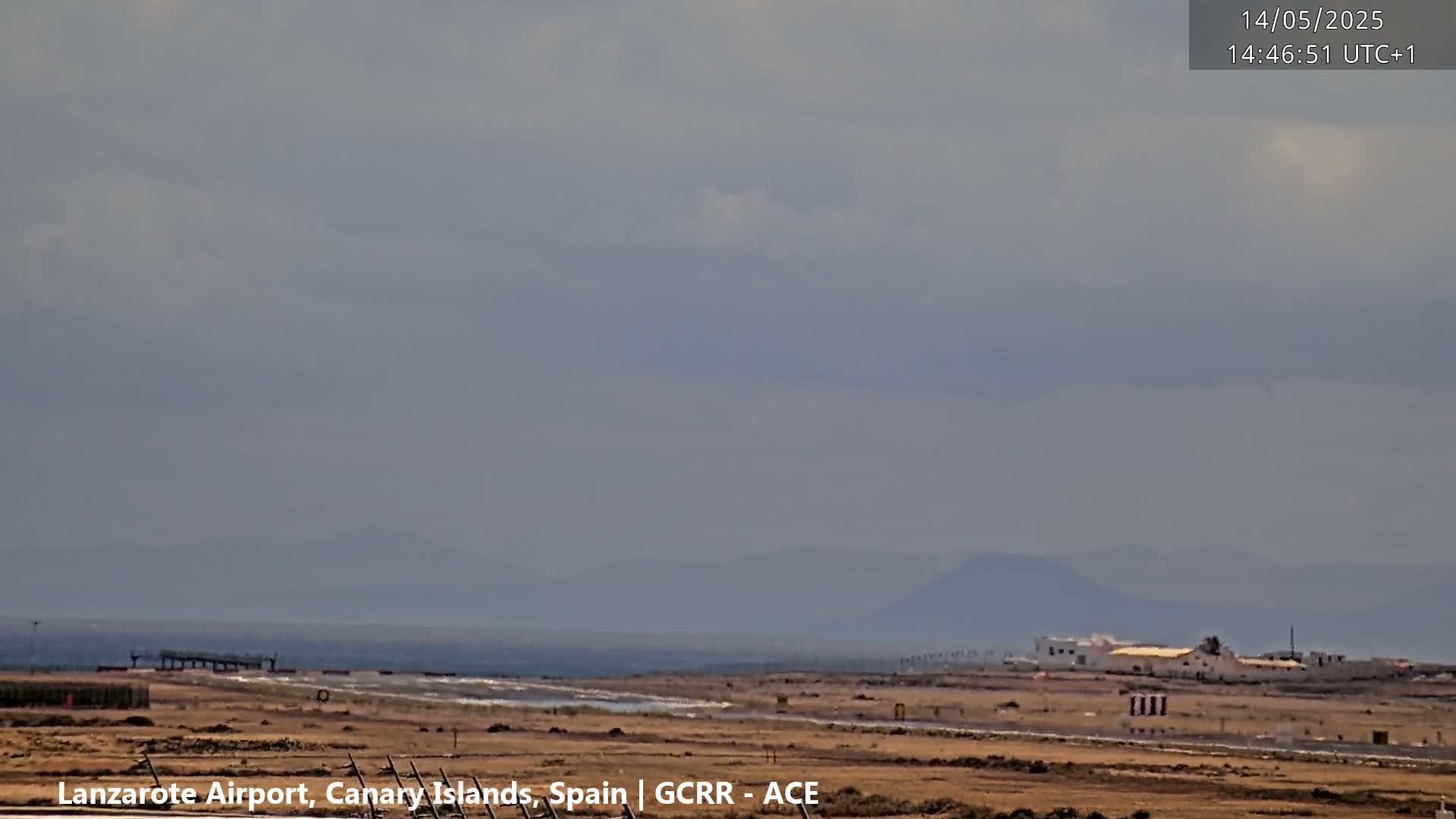 A mostly cloudy day reveals a coastal airport with a runway and a distant ocean under a hazy sky, with mountains visible in the far background.