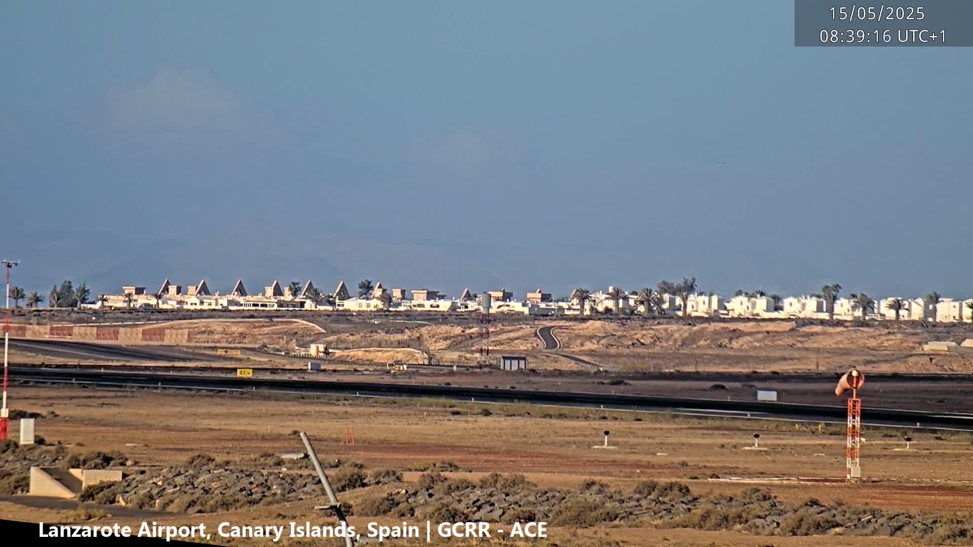 A clear, sunny day reveals a distant row of low-rise buildings and white tanks beyond an airport runway.