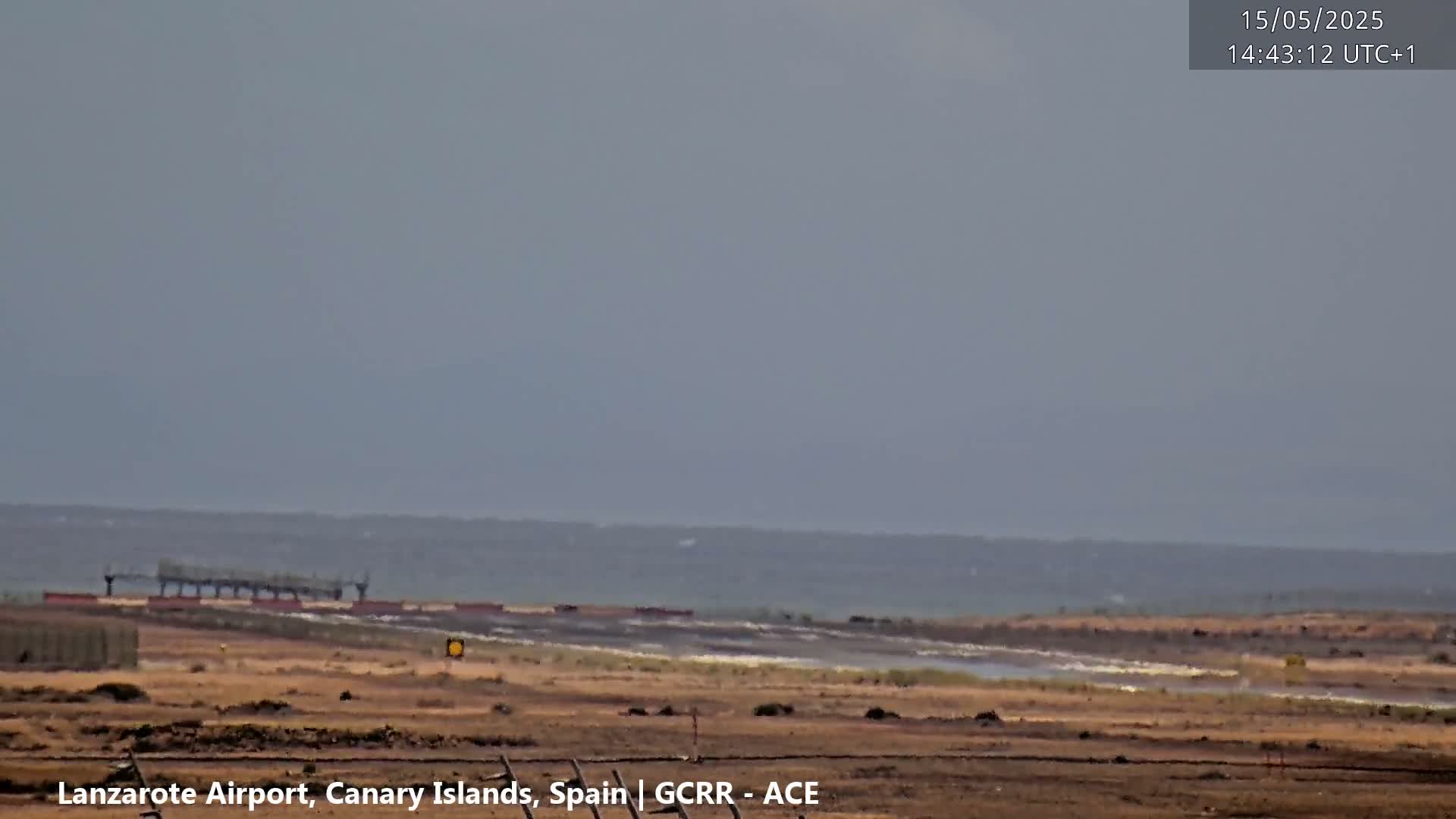 A hazy, overcast day reveals a flat, brown landscape with a distant body of water and a portion of an airport in the middle ground.