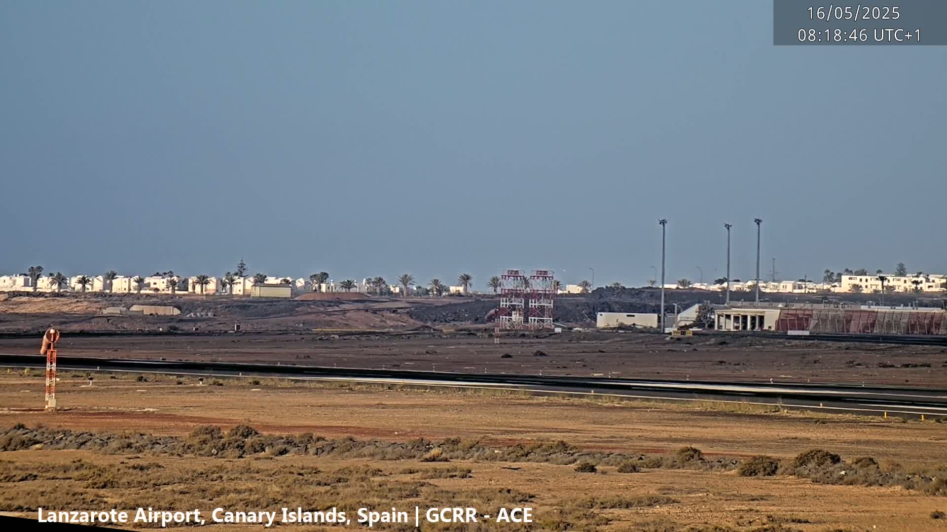 A flat, arid landscape featuring an airport runway, a distant row of buildings, and a tall metal structure under a clear, pale blue sky.