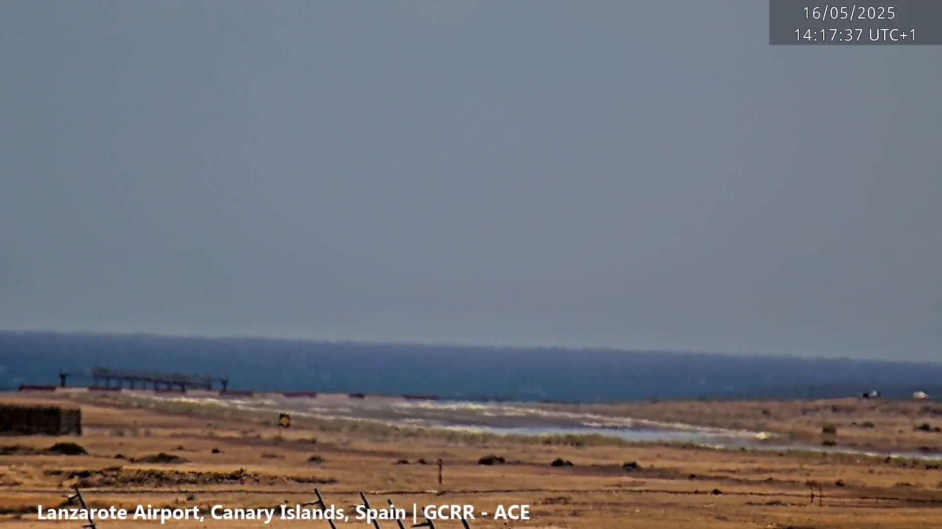 A hazy view shows a flat, dry landscape leading to a calm ocean under a light grey sky.