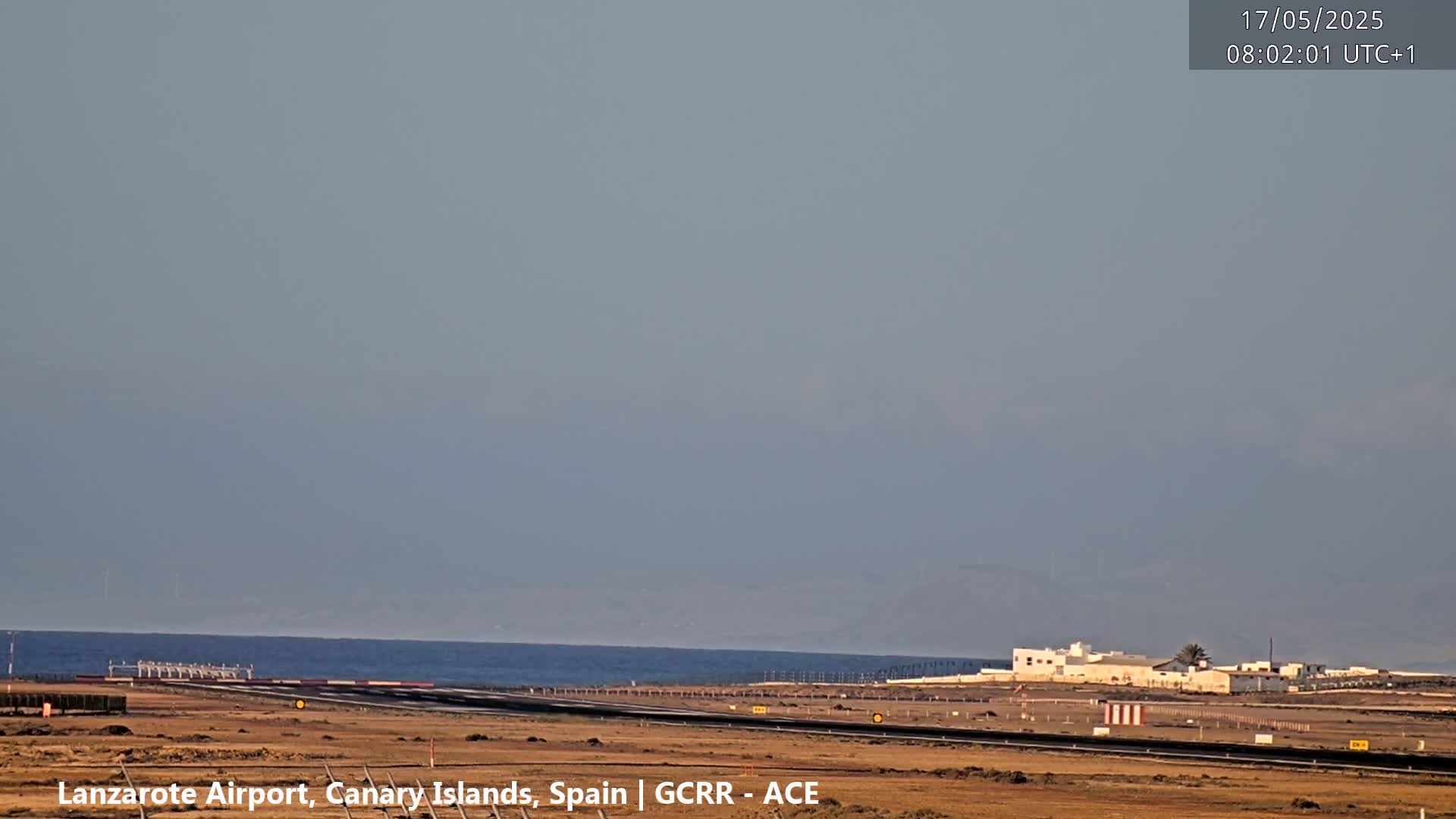 A hazy view of an airport runway and terminal building next to a calm ocean under a clear, light-grey sky.