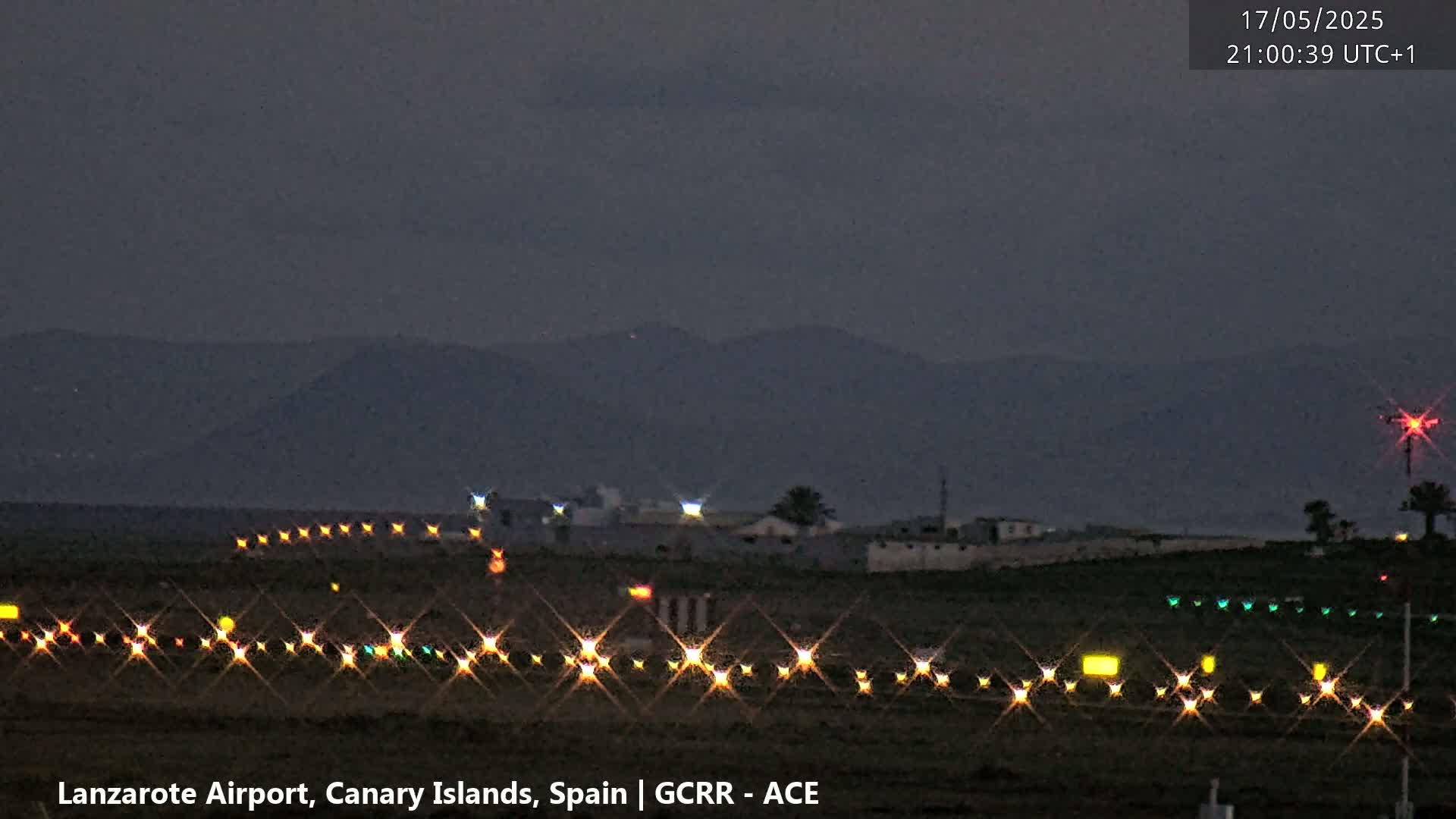 A dark, nighttime view of an airport runway with illuminated lights, bordered by low buildings and distant mountains under a dusky sky.