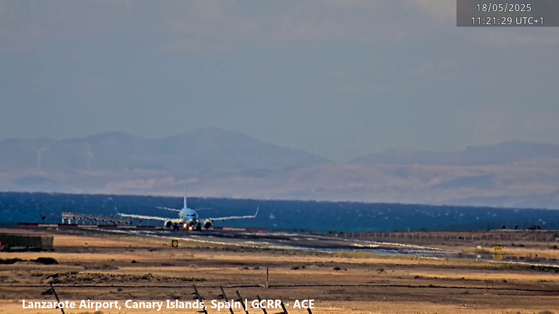 A passenger airplane taxis on a runway near a body of water under a hazy sky, with distant mountains visible in the background.