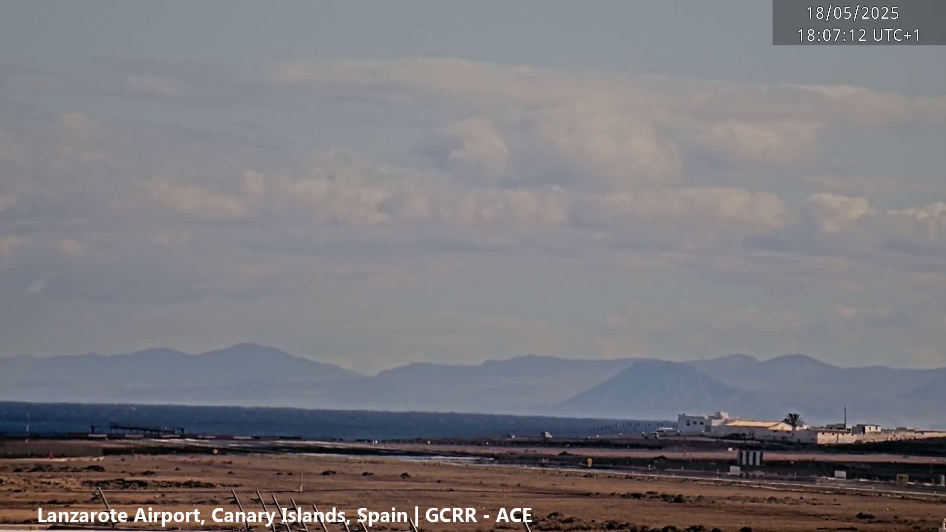 A mostly clear sky with scattered clouds hangs over a flat, brown landscape featuring a distant mountain range and a body of water.