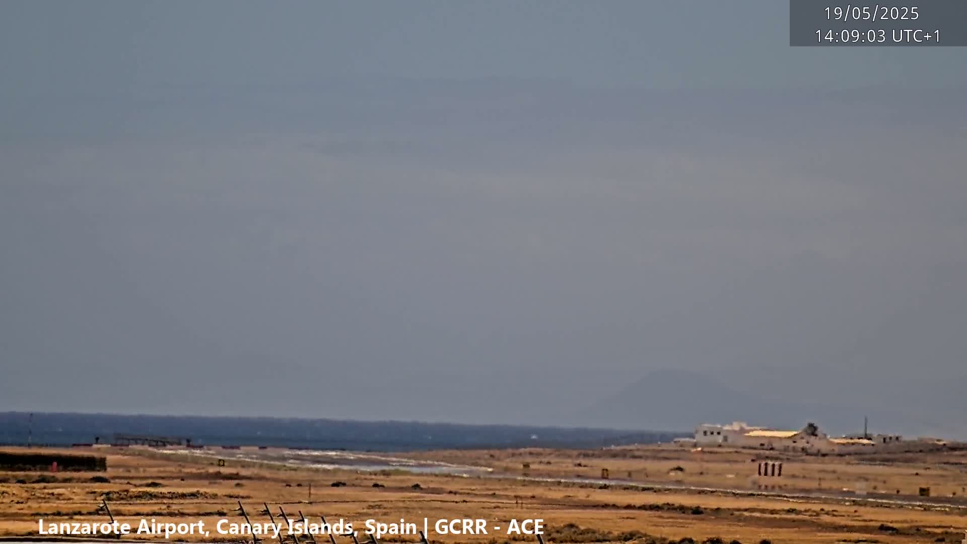 A hazy, overcast day shows a coastal airport with a runway, buildings, and a calm ocean in the distance.
