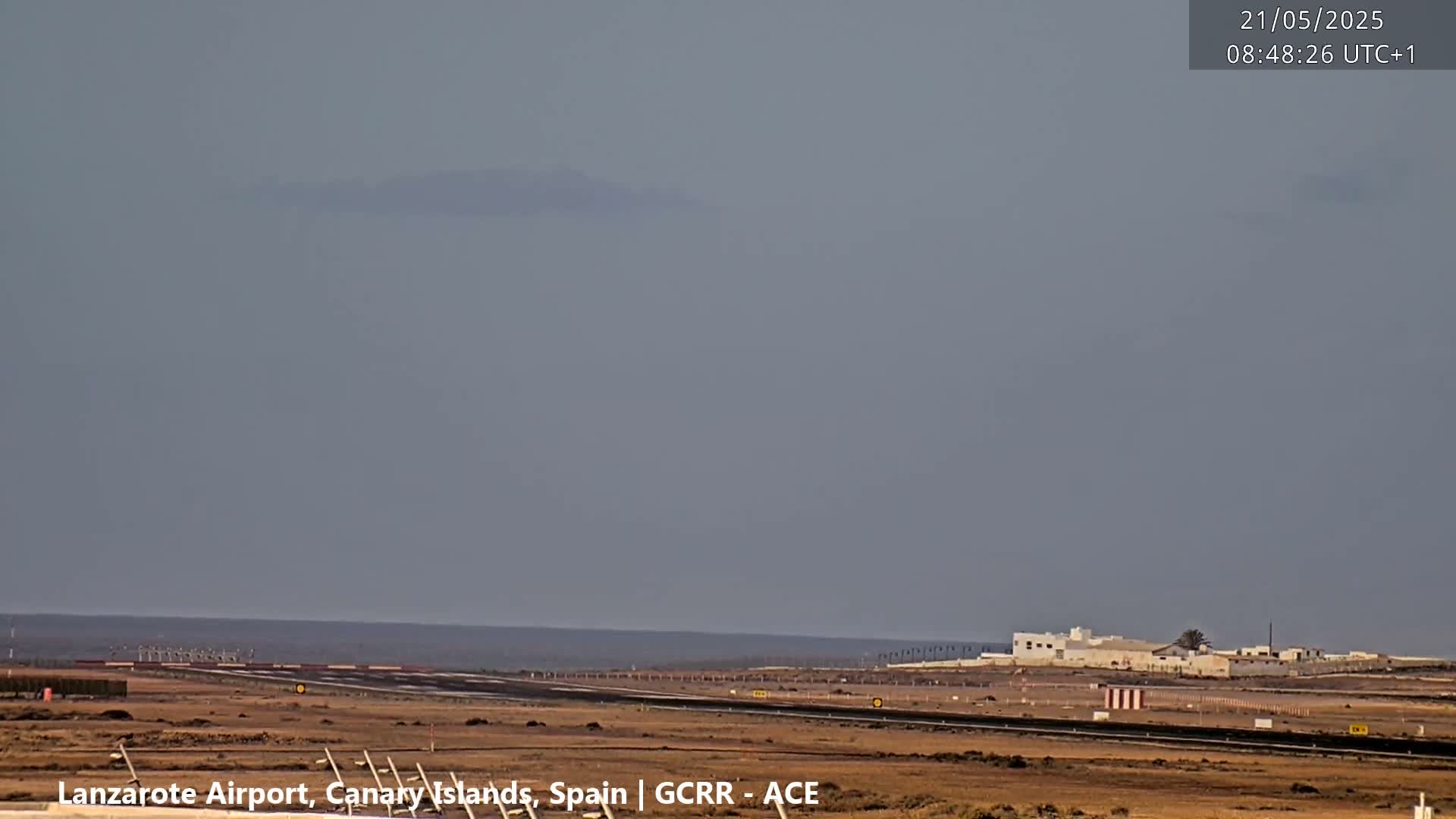 An airport runway and terminal buildings are visible on a hazy day, with a calm ocean in the background.