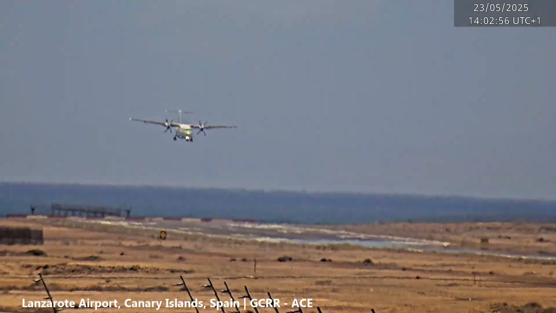 A turboprop airplane is approaching an airport runway on a hazy day over a flat, arid landscape with a body of water in the distance.