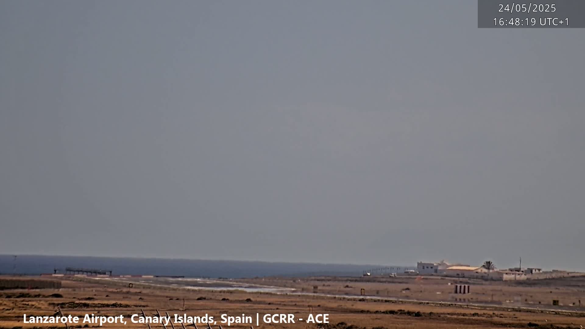 A hazy, sunny day overlooks a flat, desert-like airport runway bordering a calm ocean.