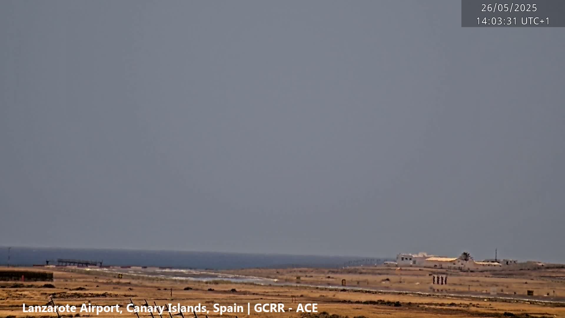 A hazy, overcast sky overlooks a flat, arid landscape with a distant ocean, a small building, and a runway.
