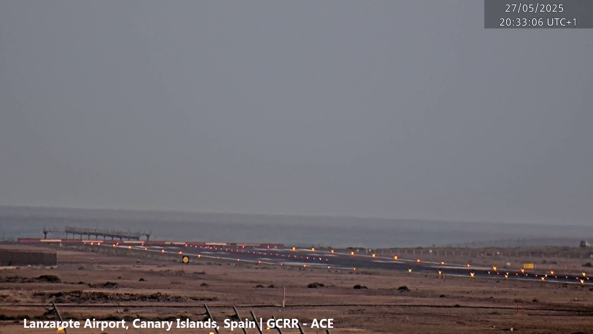 An airport runway with illuminated lights extends toward a calm ocean under a gray, overcast sky.