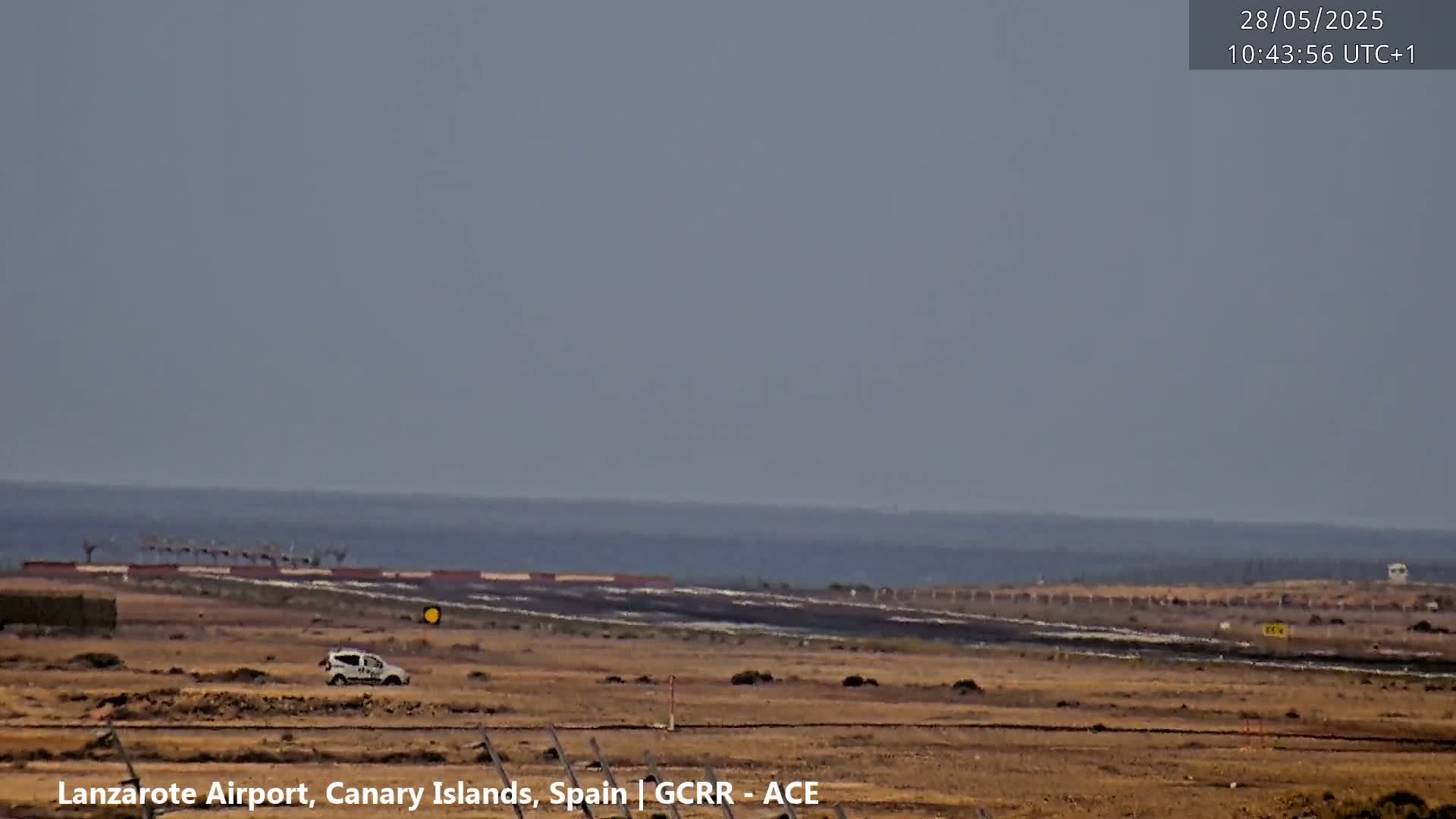A light-colored vehicle is parked on a flat, barren expanse of land near a runway that overlooks a calm ocean under a hazy sky.