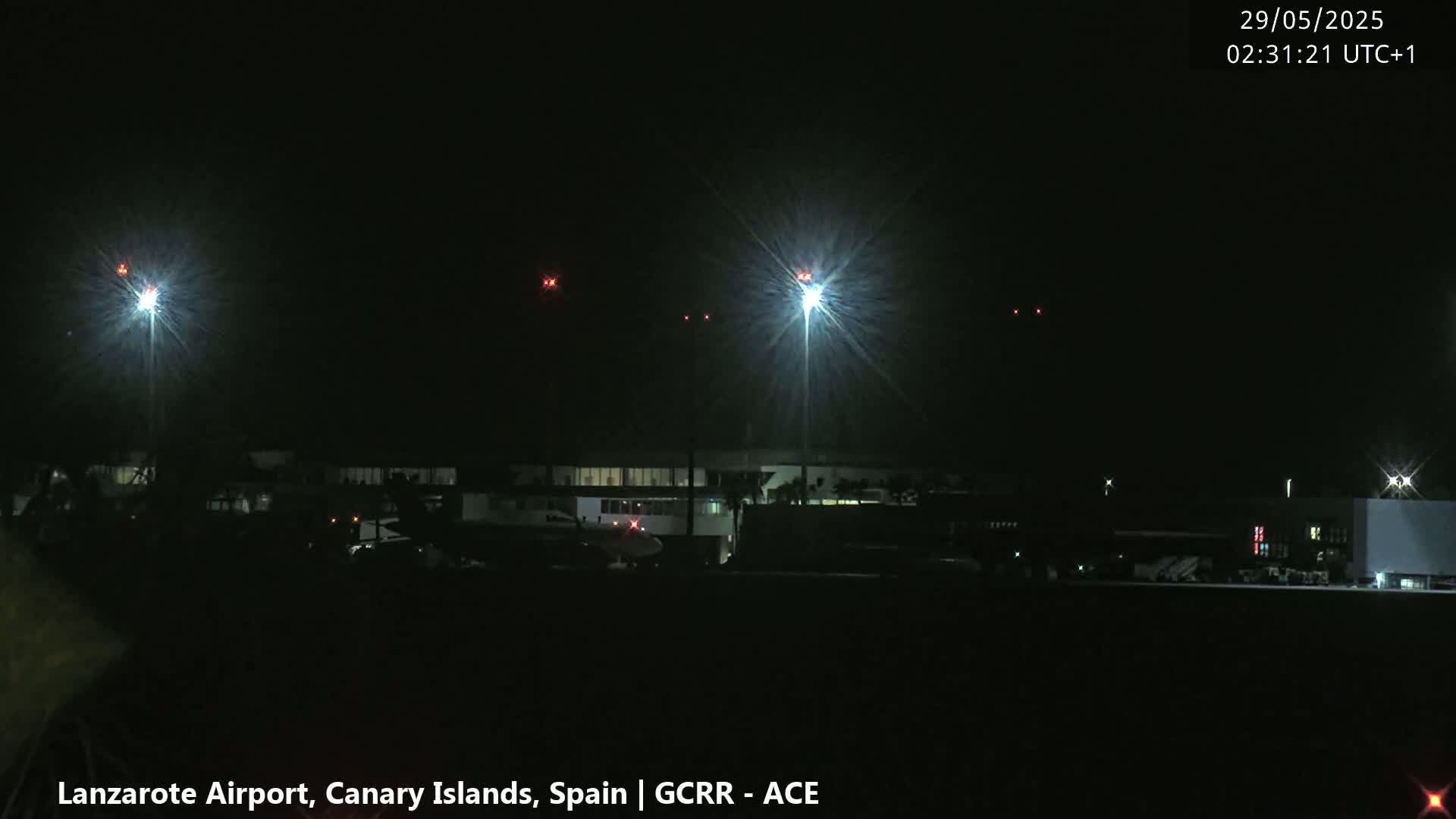 At night, brightly lit airport buildings and tarmac are visible under a dark sky, with several distant red lights possibly indicating aircraft.