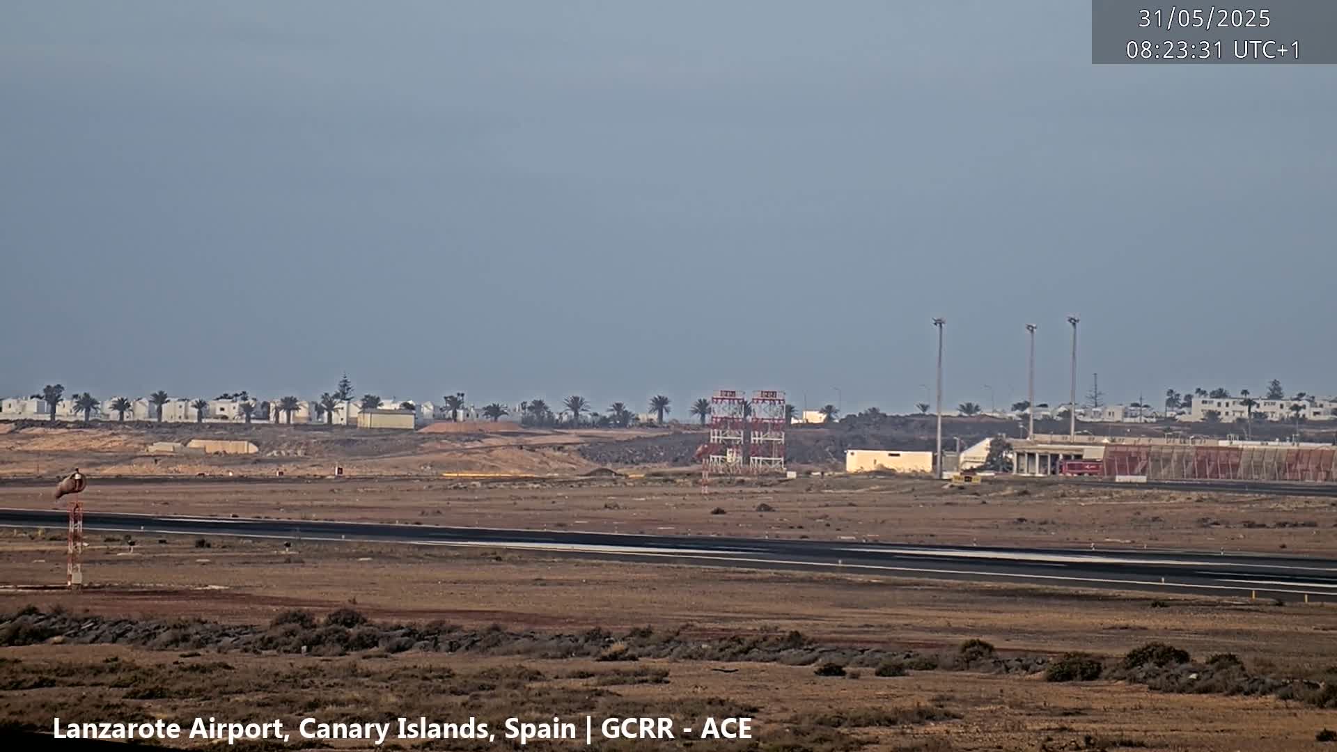 A flat, arid landscape with a runway, airport infrastructure, and distant buildings under a clear, pale sky.
