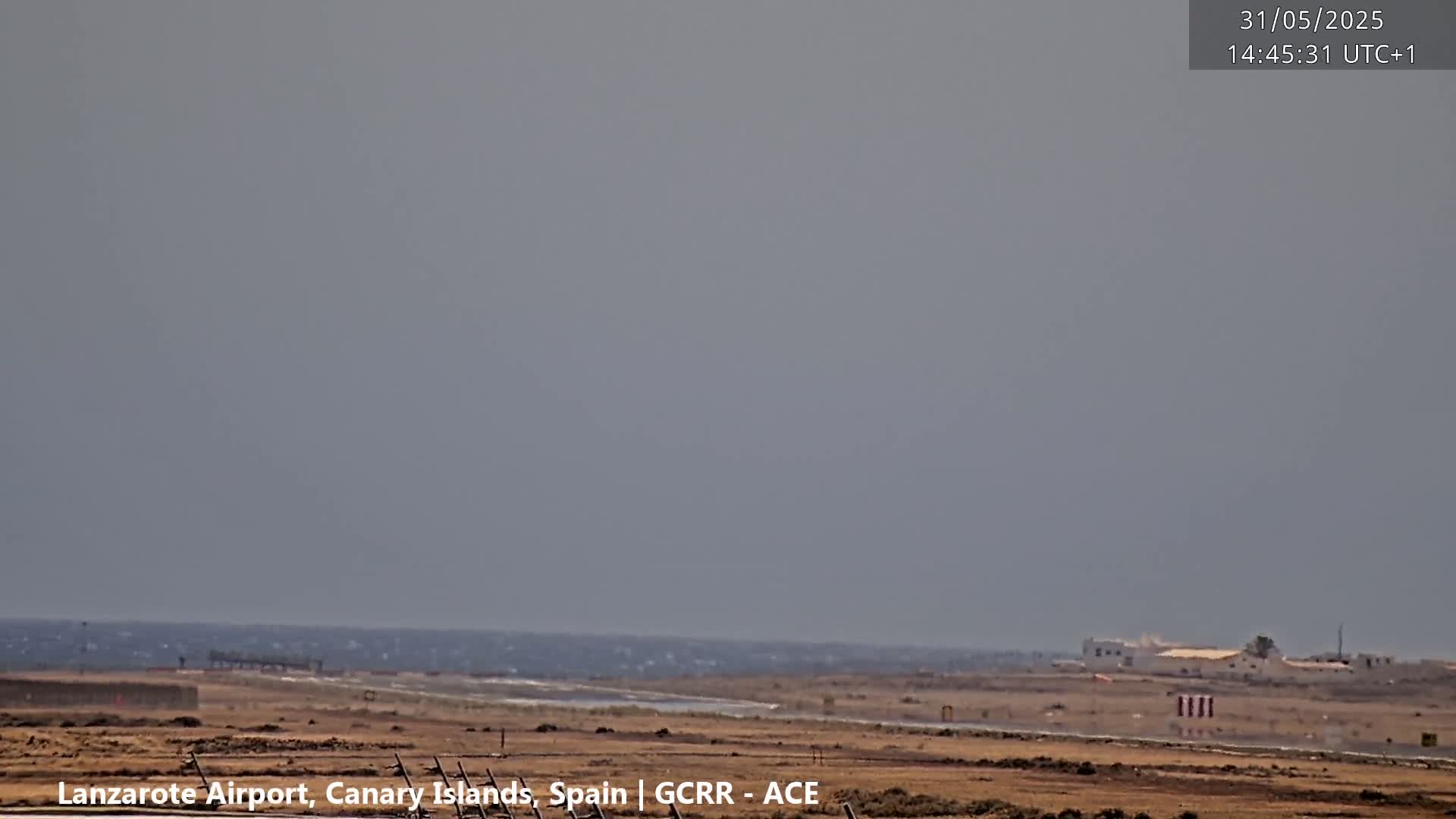 A hazy, overcast day shows a flat, arid landscape with a body of choppy water in the distance, a runway, and a few buildings.