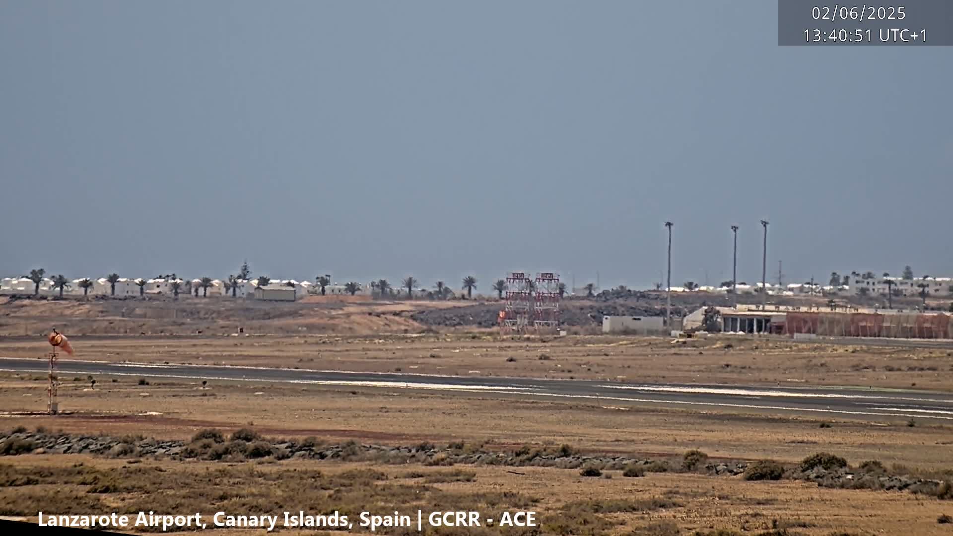 A mostly clear day shows an airport runway and windsock, with low-lying buildings and sparse vegetation visible in the distance.