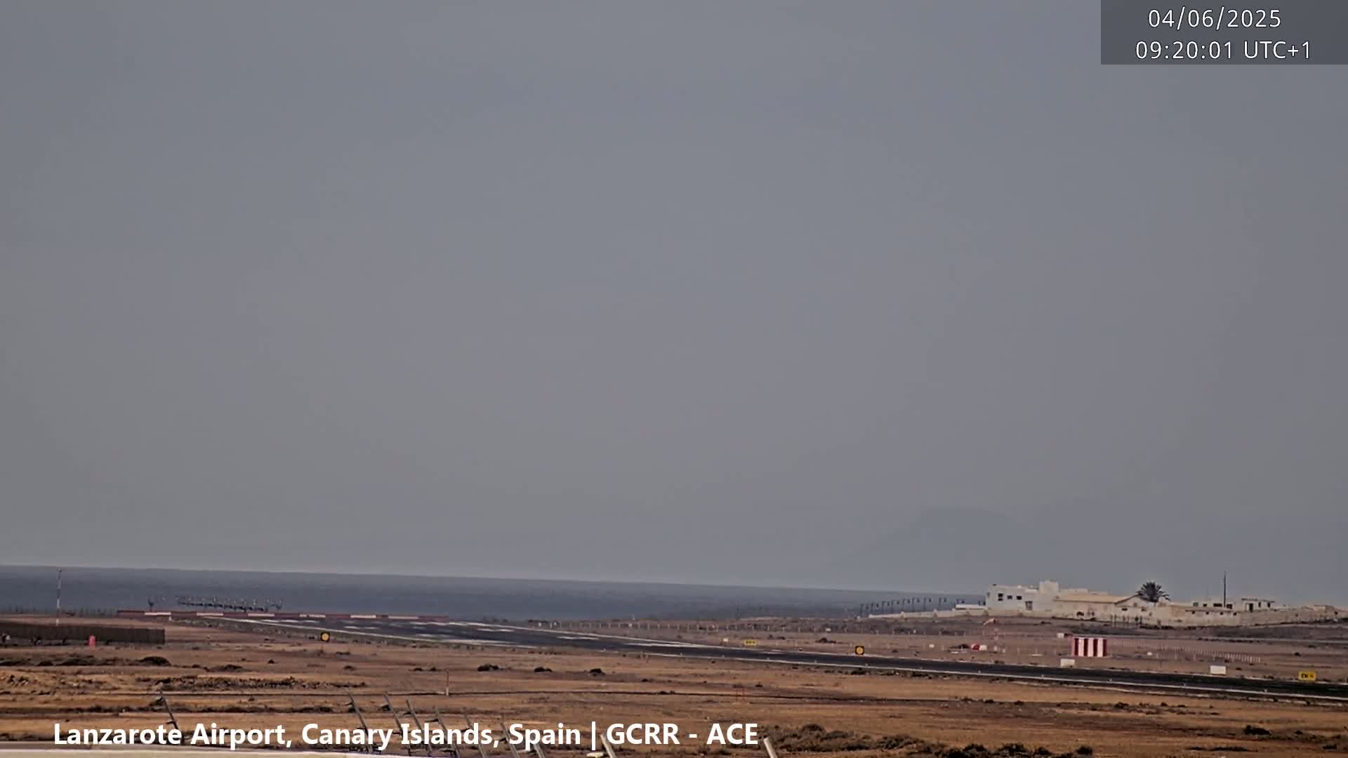 A hazy, overcast day reveals a runway leading to a calm ocean, with a small cluster of buildings visible in the distance.