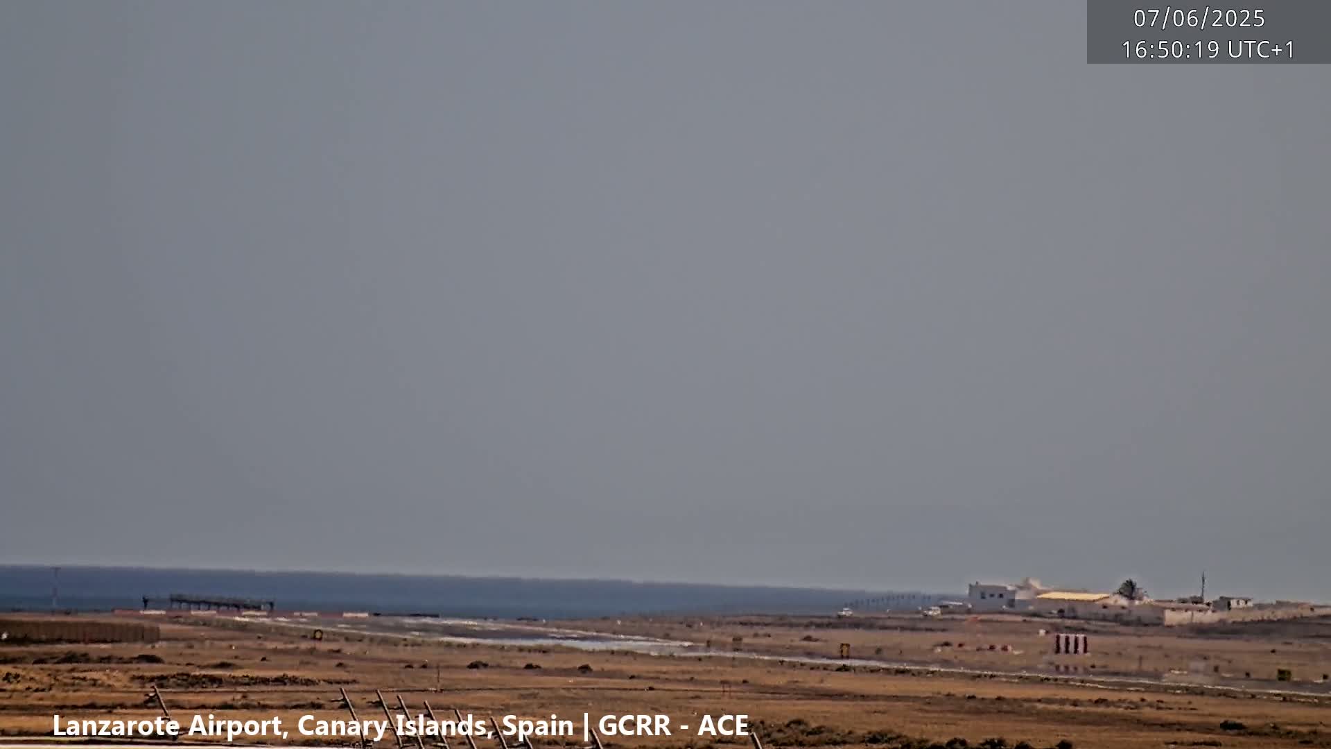A hazy, flat landscape shows a calm ocean in the distance, a runway, and sparse, low-lying buildings under a light-grey sky.