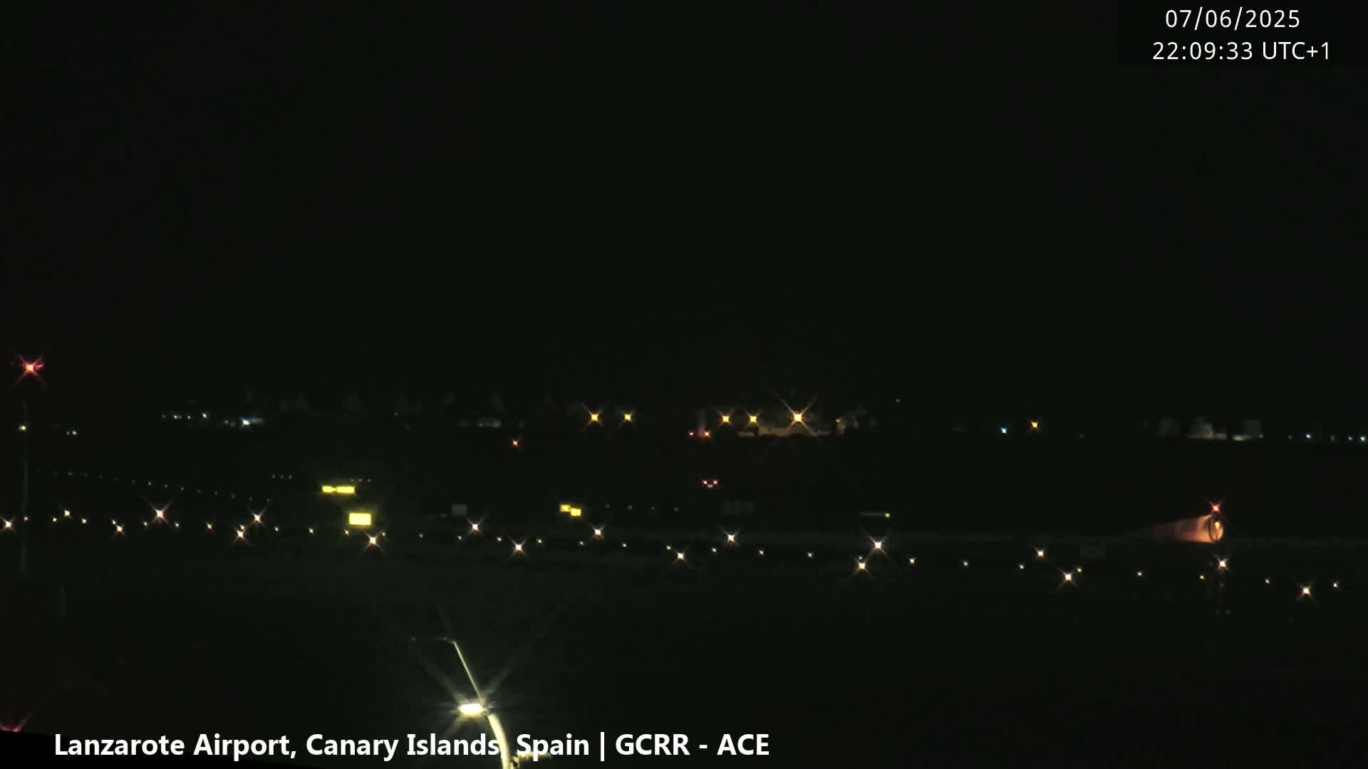 A nighttime view of an airport runway with dimly lit ground vehicles and distant building lights under a clear, dark sky.